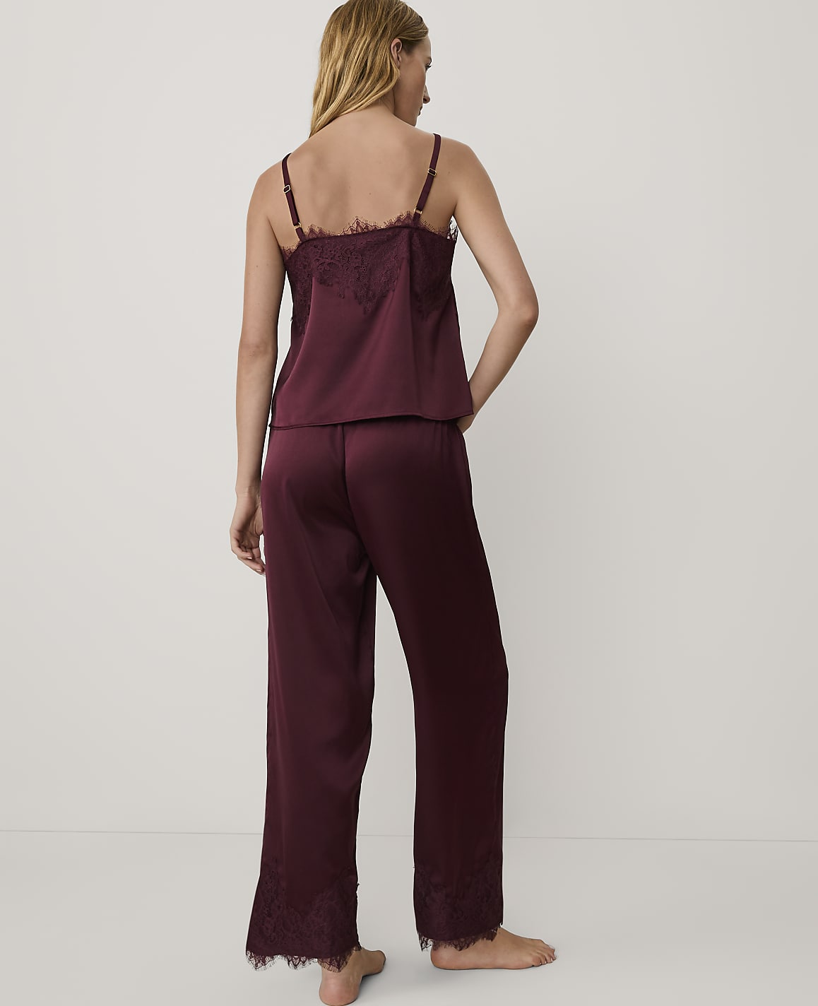 A woman stands with her back to the camera wearing burgundy satin pajamas with lace trim against a plain white background.