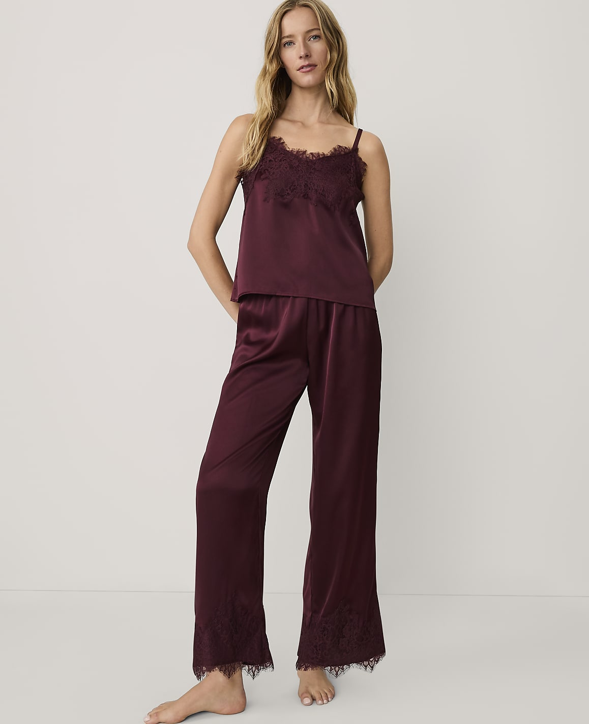 A woman wearing burgundy satin pajamas with lace trim stands against a plain white background.