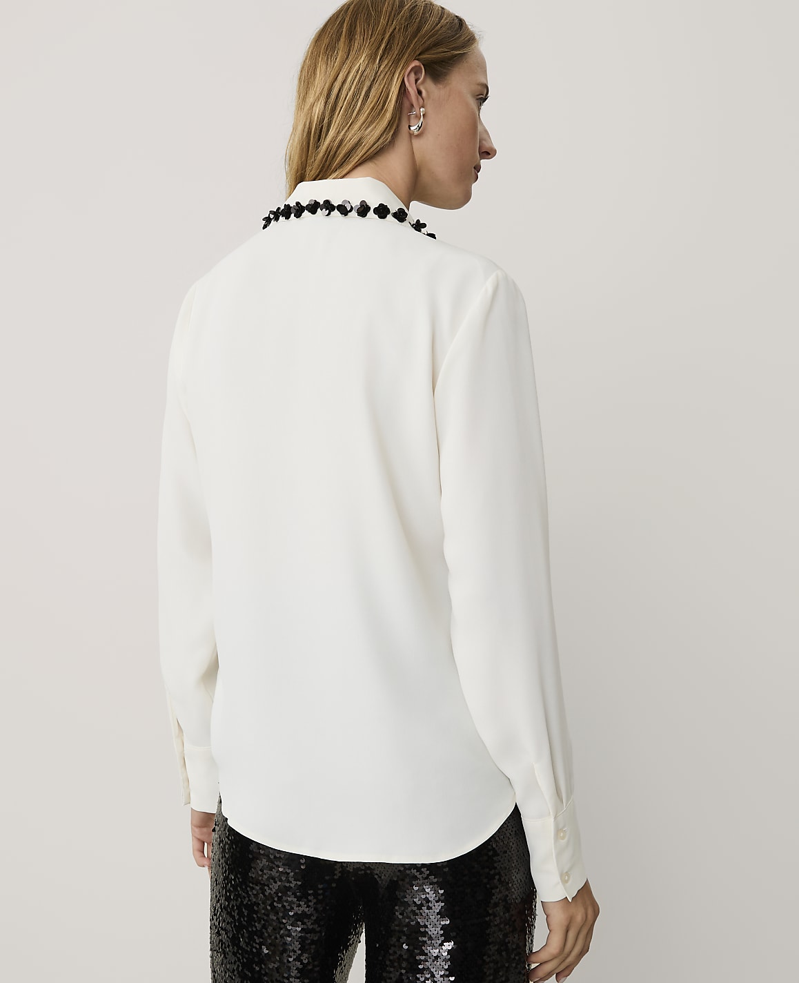 A woman wearing a white long-sleeved blouse with a beaded collar and black sequined pants stands against a plain light gray background.