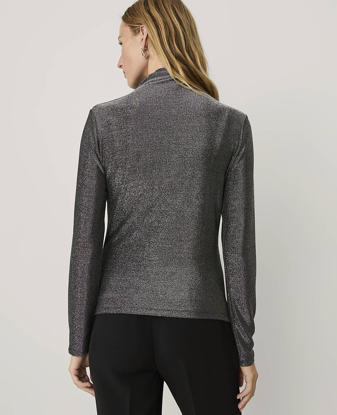 A woman wearing a shimmering grey long-sleeved top and black trousers stands with her back to the camera against a plain white background.