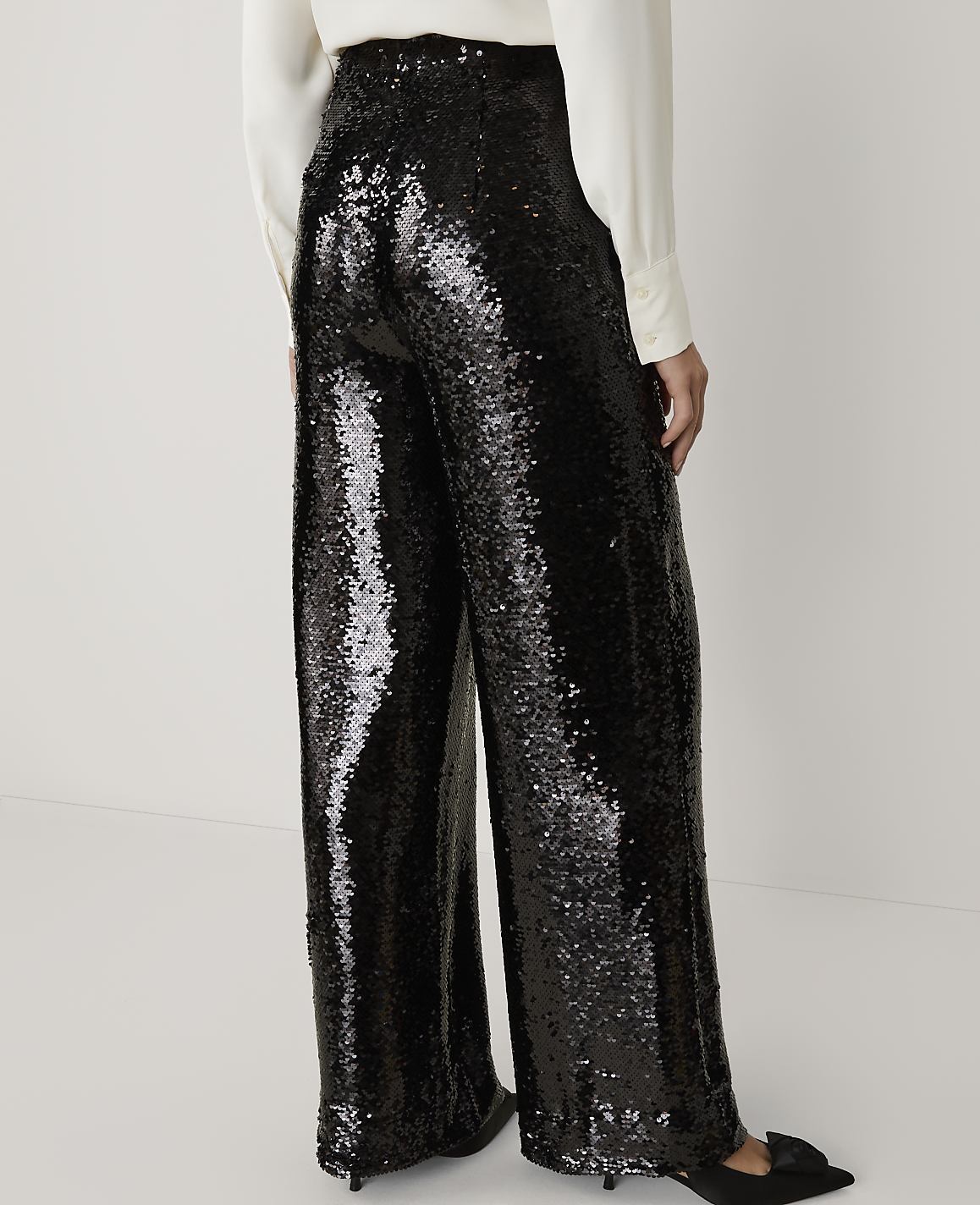 A person wears shimmering black sequined wide-leg trousers and a white blouse against a plain, light-colored background.