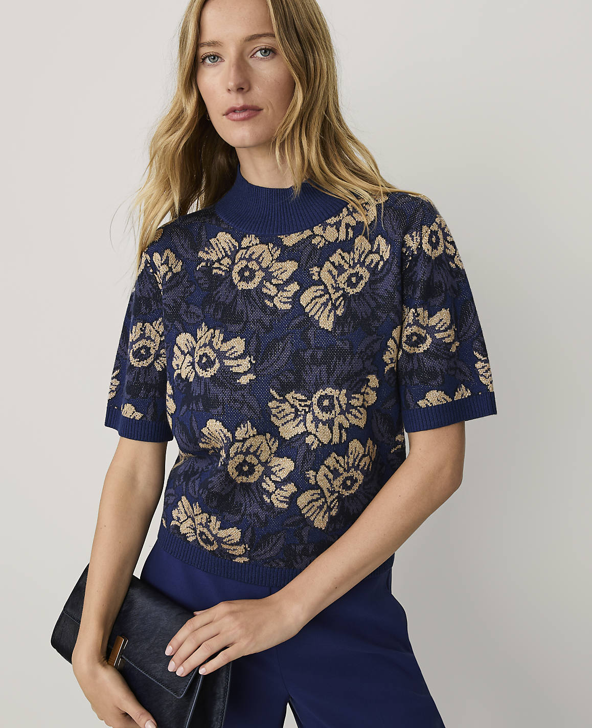 A blonde woman wearing a blue floral knit top and matching trousers holds a black clutch against a plain light grey background.