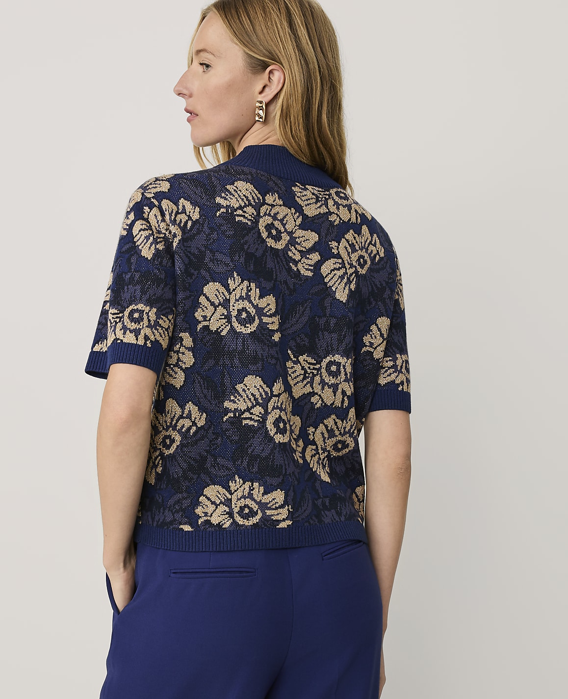 A woman wearing a blue and gold floral knit top and blue trousers stands against a plain light gray background.