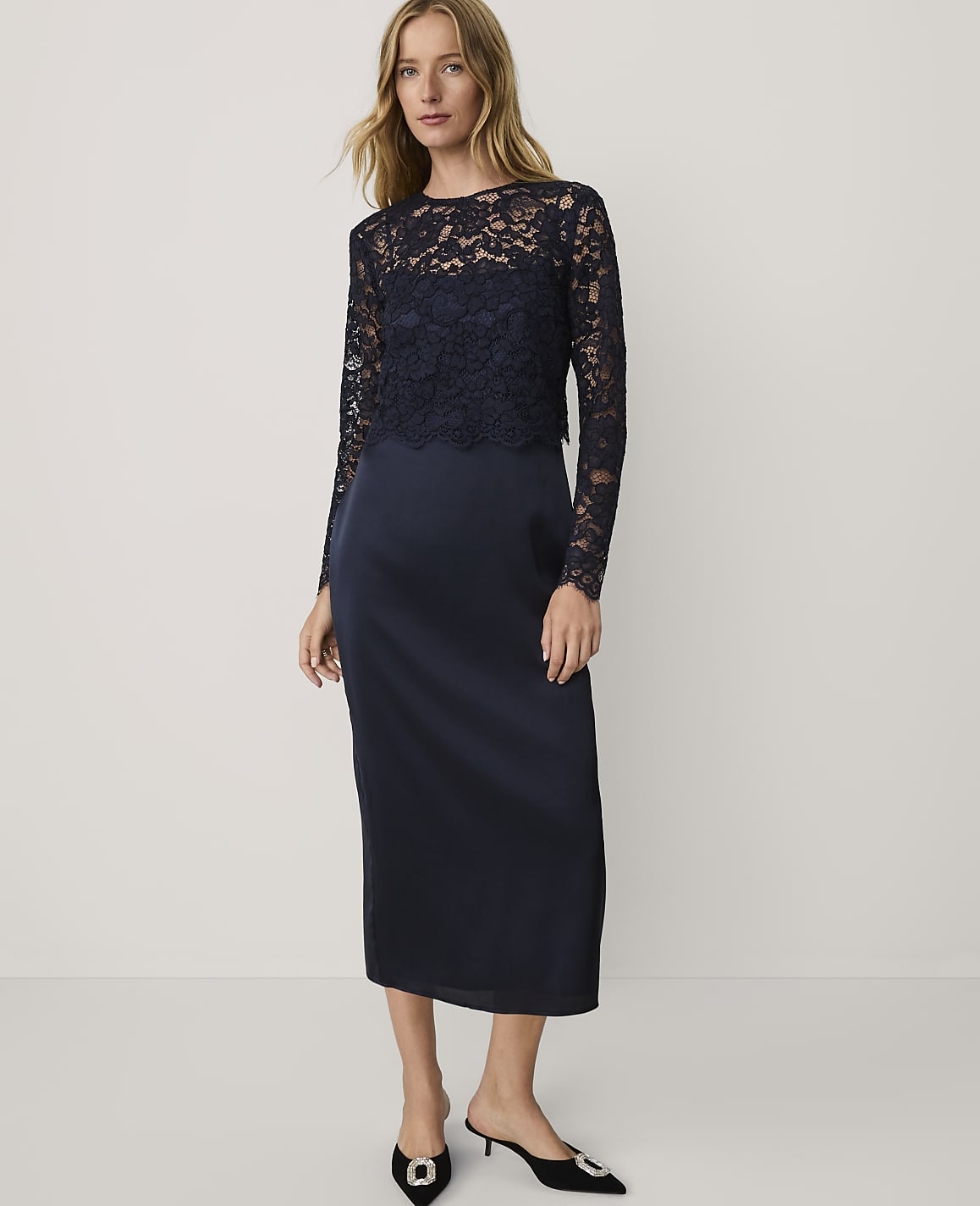 A woman wearing a navy blue lace and satin dress and black heels stands against a plain white background.