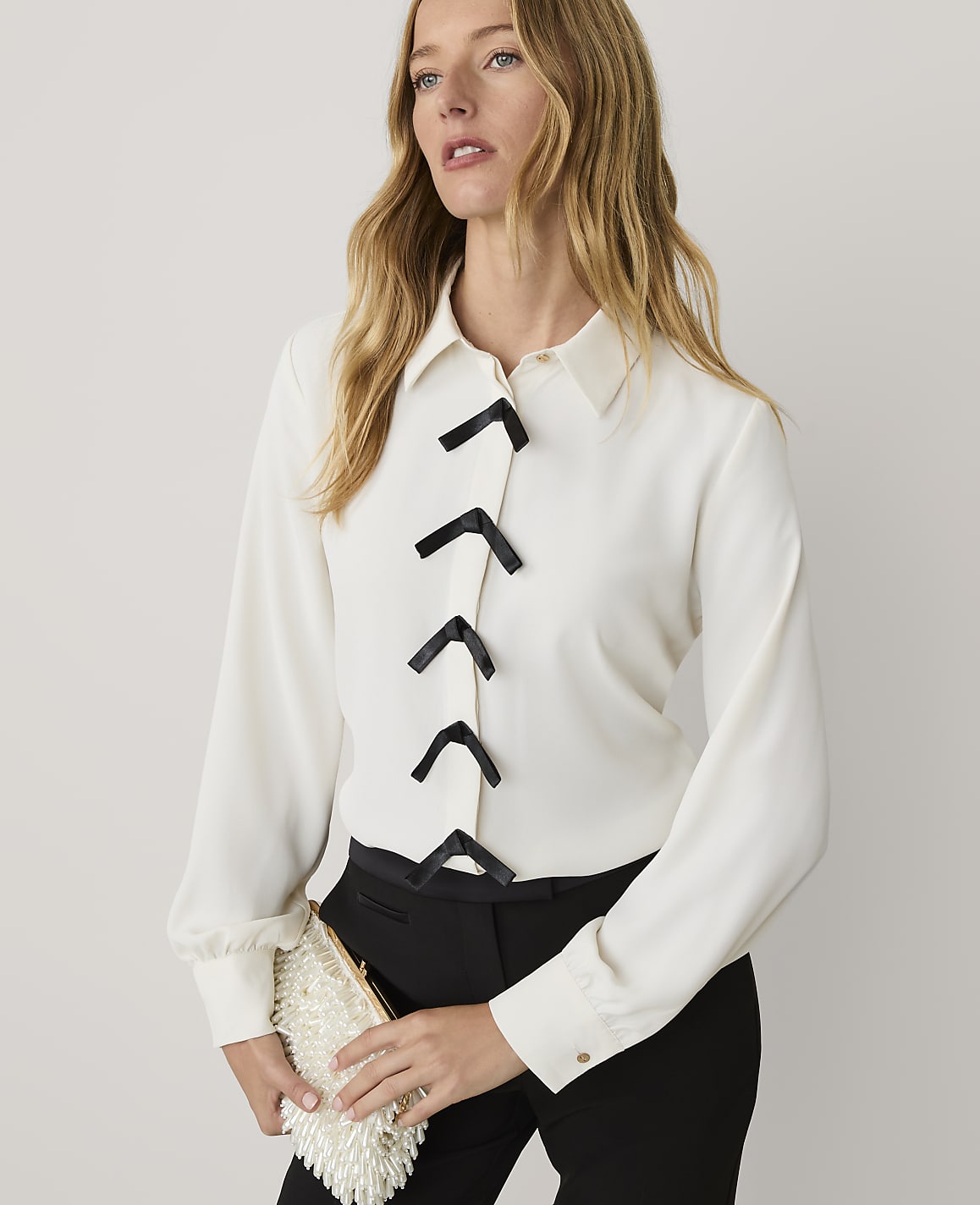 A blonde woman wearing a white blouse with black accents and black trousers stands against a plain white background.