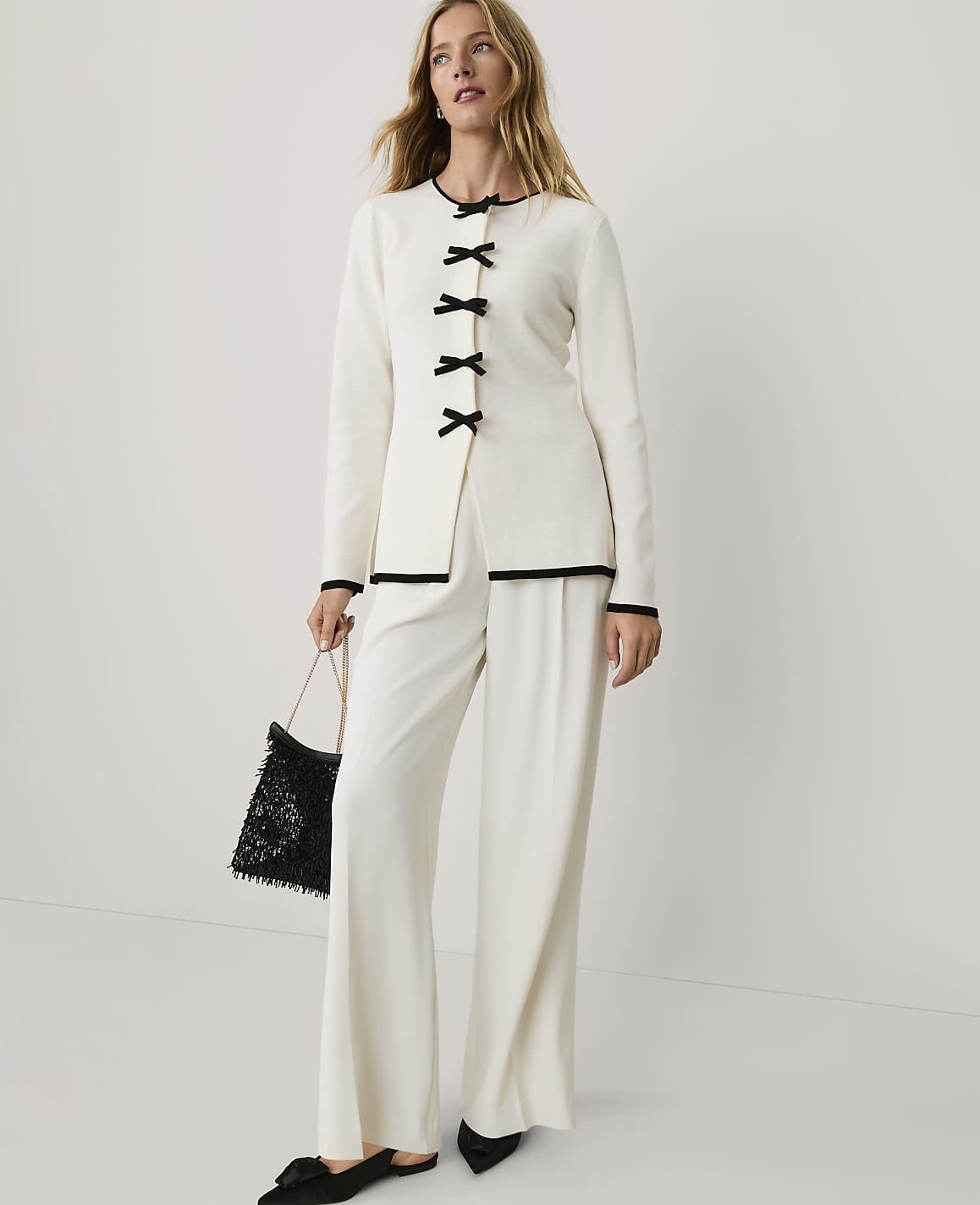 A woman wearing a white two-piece outfit with black accents and holding a black bag stands against a plain white background.