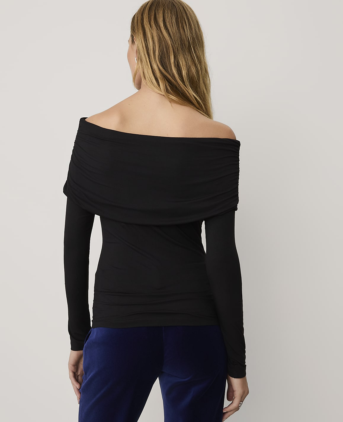 A woman wearing a black off-the-shoulder top and blue velvet pants stands against a plain light gray background.