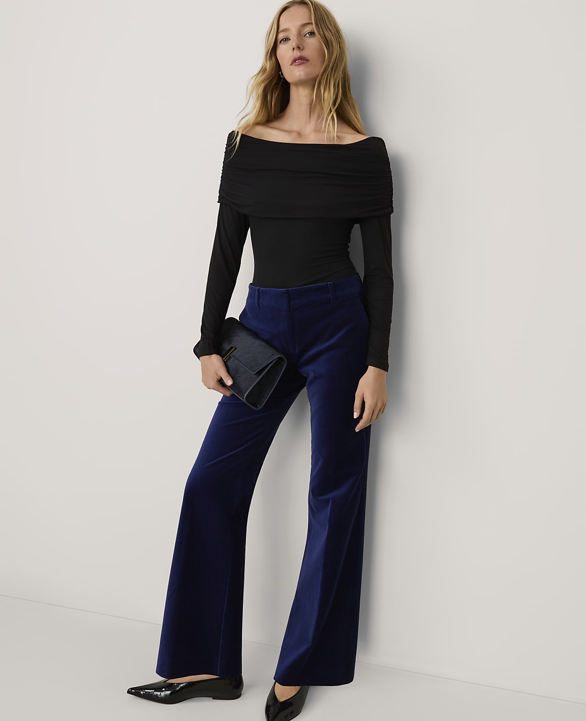 A woman wearing a black off-the-shoulder top and wide-leg blue velvet trousers stands against a plain white wall.