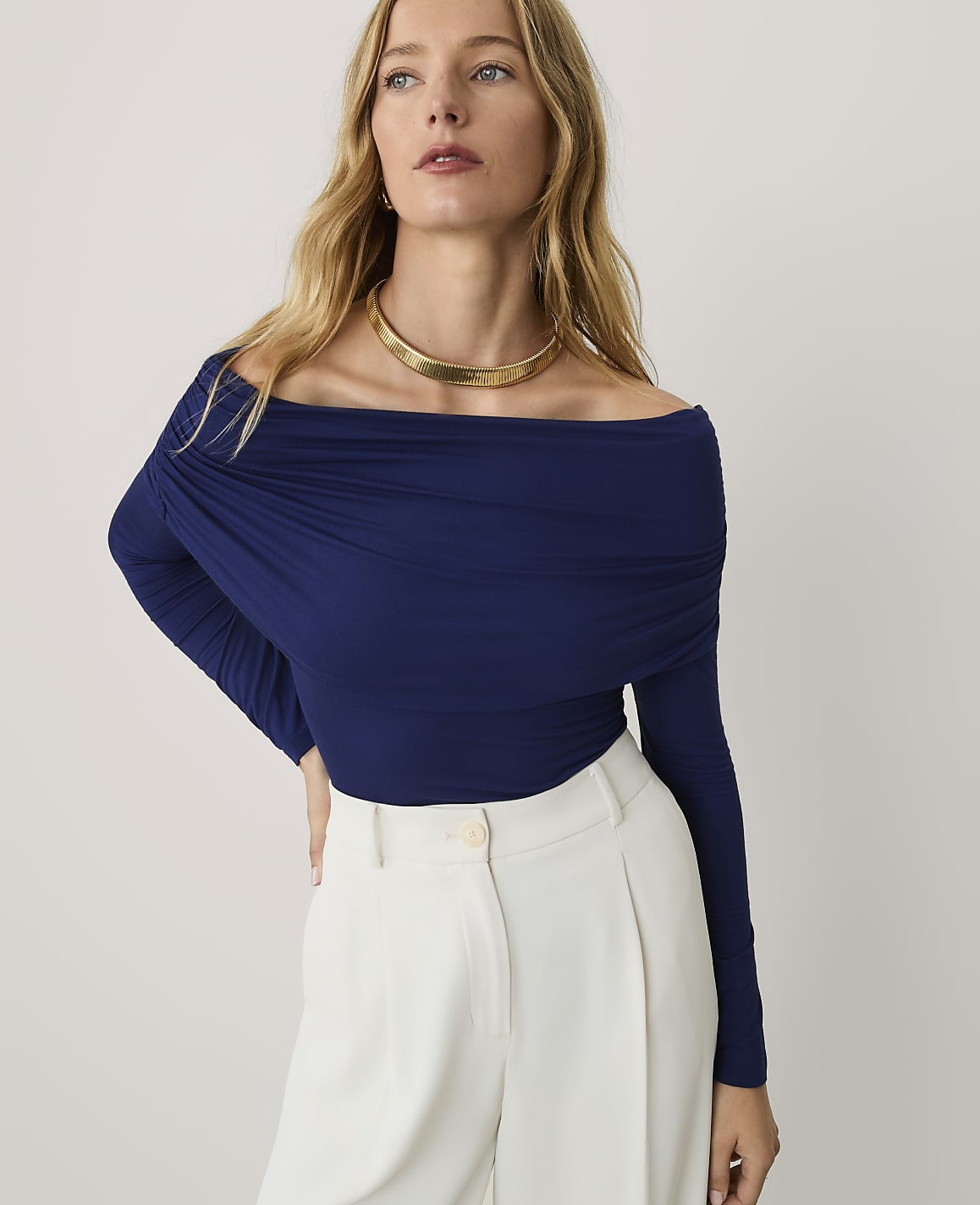 A blonde woman wearing a navy blue off-the-shoulder top and white trousers stands against a plain light gray background.