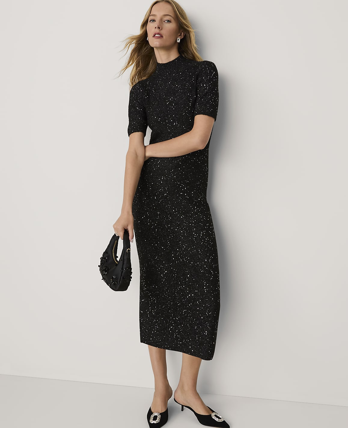 A woman wearing a sparkling black midi dress and black heels stands against a plain white background.