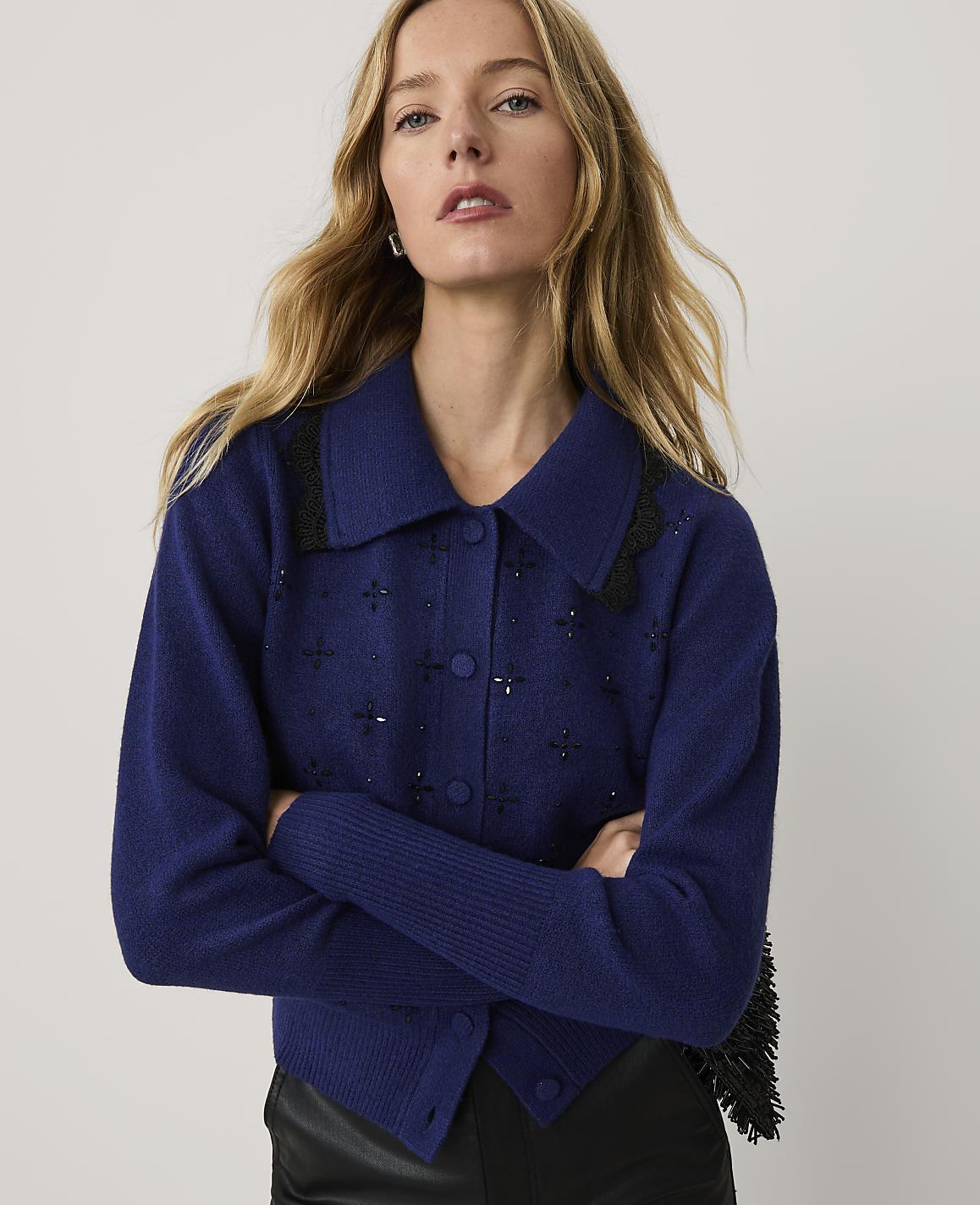 A blonde woman wearing a blue embellished cardigan and black leather pants stands against a plain white background.