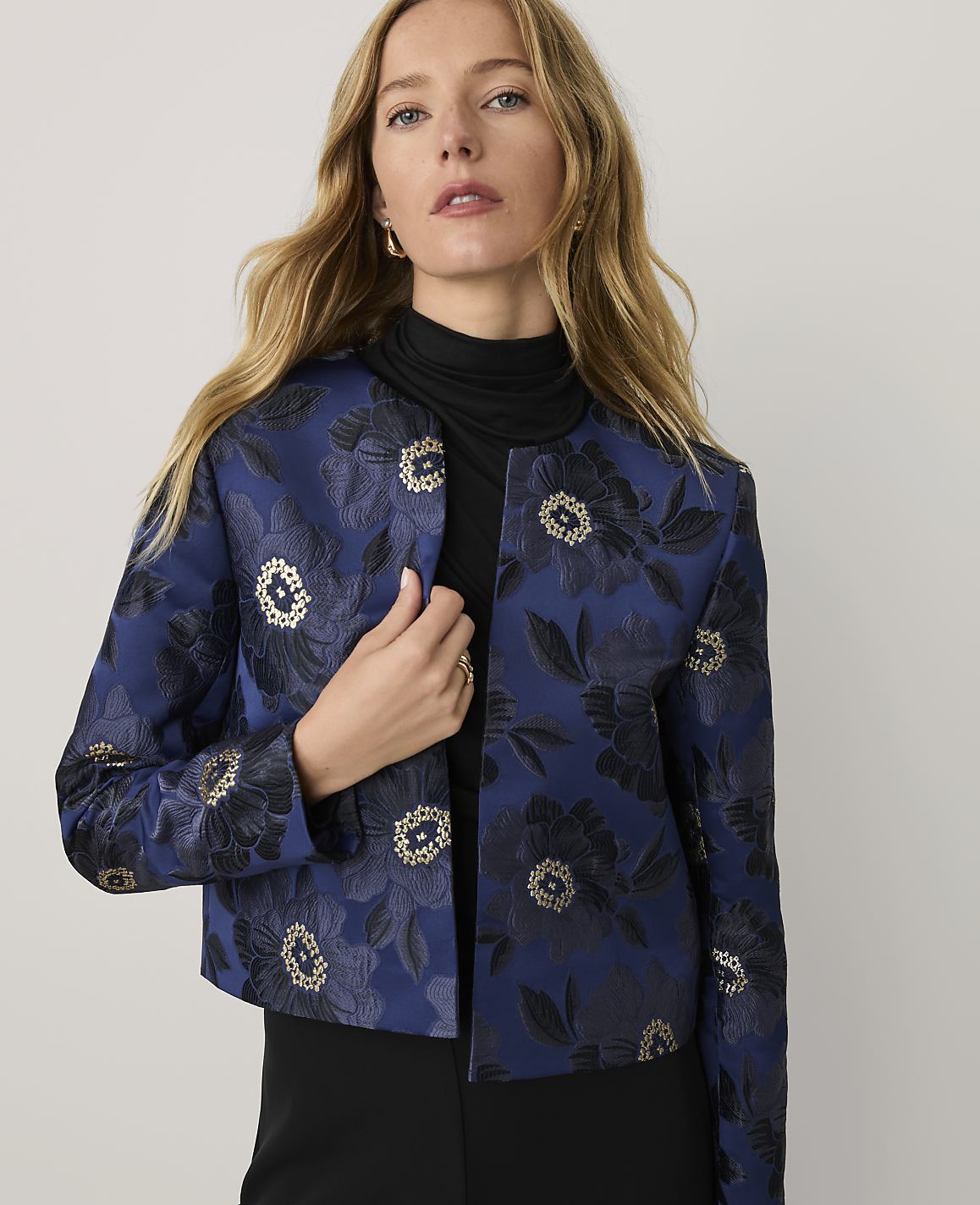 A blonde woman wearing a blue floral jacket and a black turtleneck stands against a plain white background.