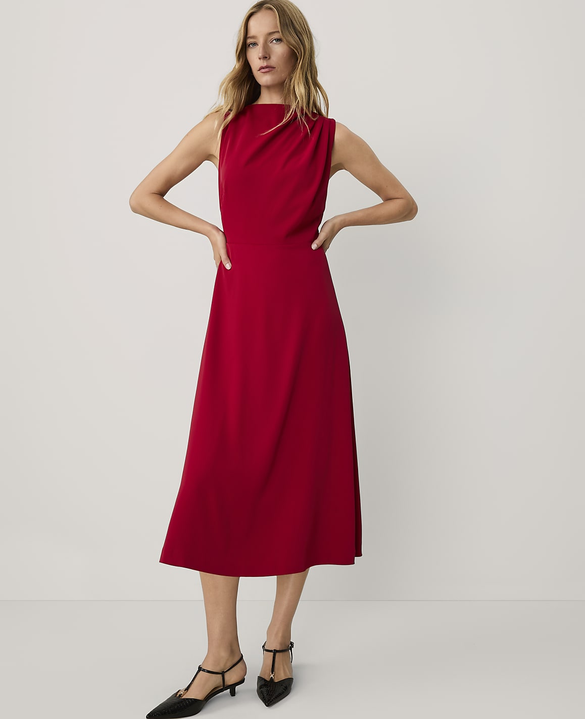 A woman wearing a sleeveless red midi dress and black pointed-toe heels stands against a plain, light-colored background.