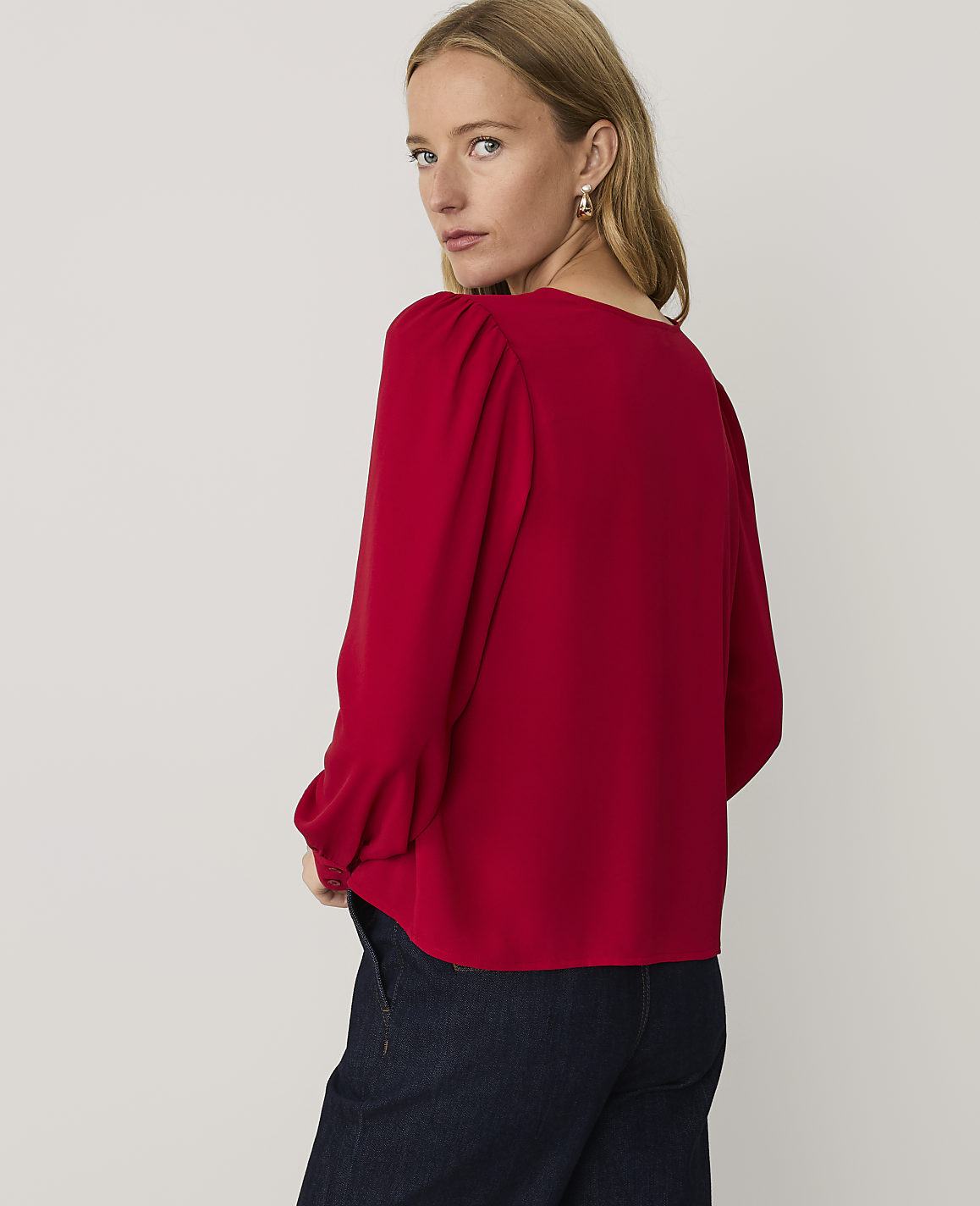 A woman wearing a red long-sleeved blouse and dark blue jeans stands against a plain, light-colored background.