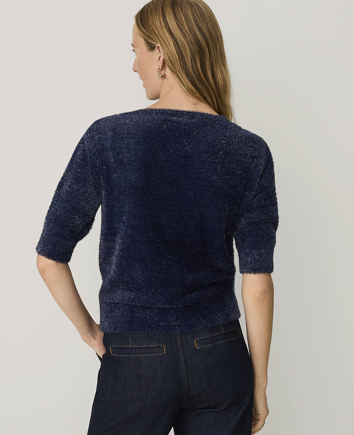 A woman wearing a fuzzy navy blue sweater and dark jeans stands with her back to the camera against a plain light gray background.