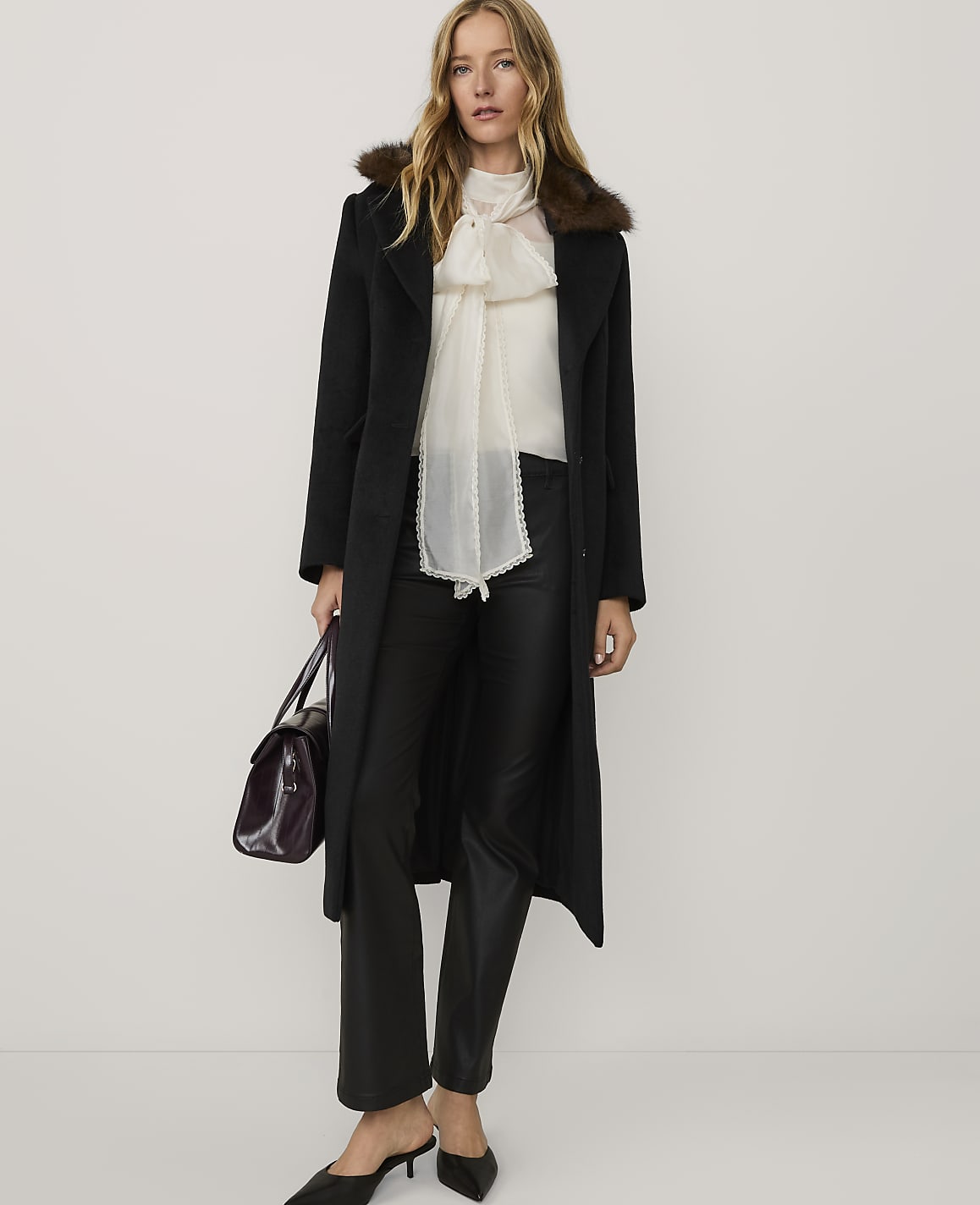 A woman wearing a black coat with a fur collar, a white blouse, and black flared trousers stands against a plain white background.