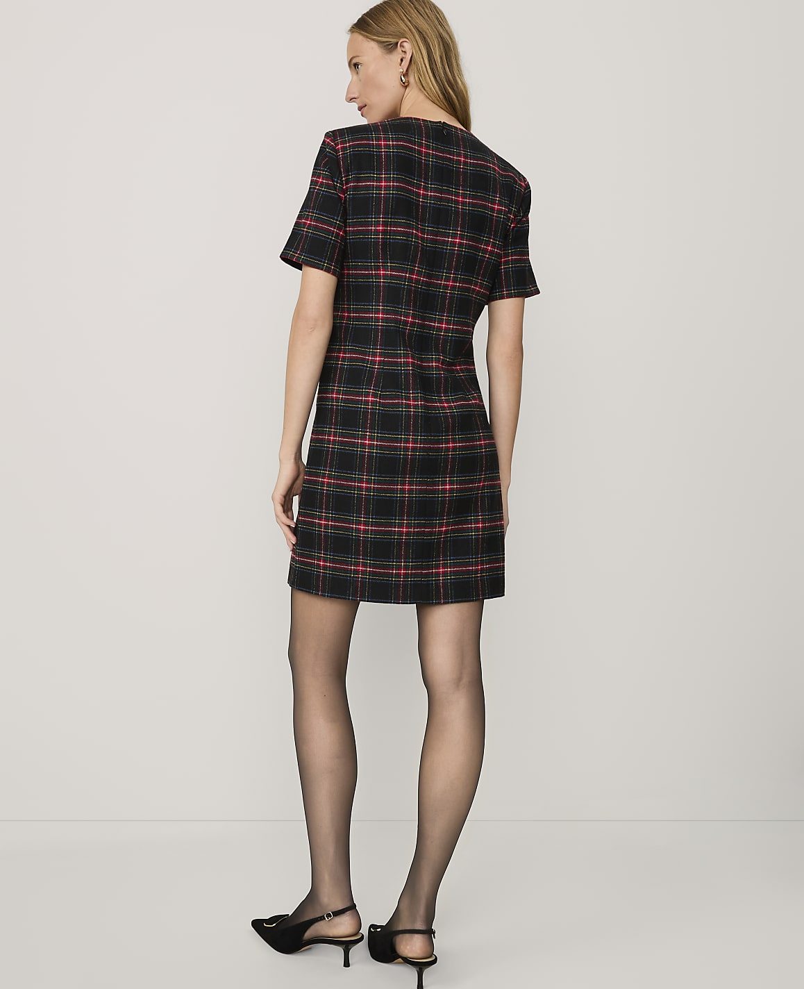 A woman wearing a red and black plaid dress, black tights, and black heels stands against a plain white background.