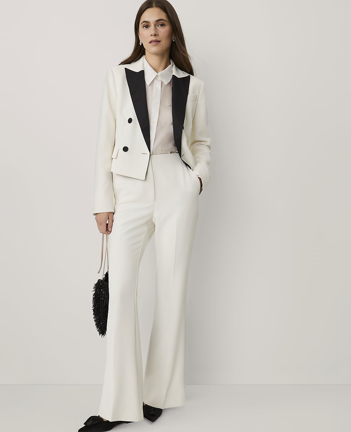 A woman wearing a white tuxedo suit with black lapels stands against a plain, light-colored background.