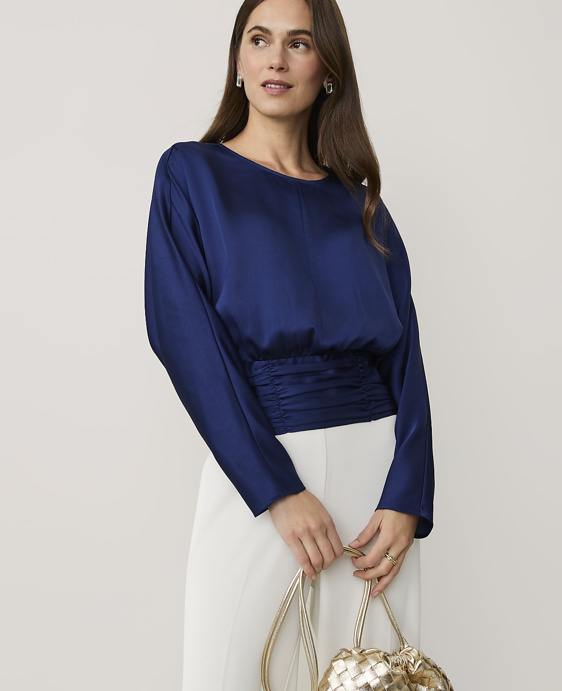 A woman wearing a navy blue satin top and white trousers holds a gold woven bag against a plain light grey background.