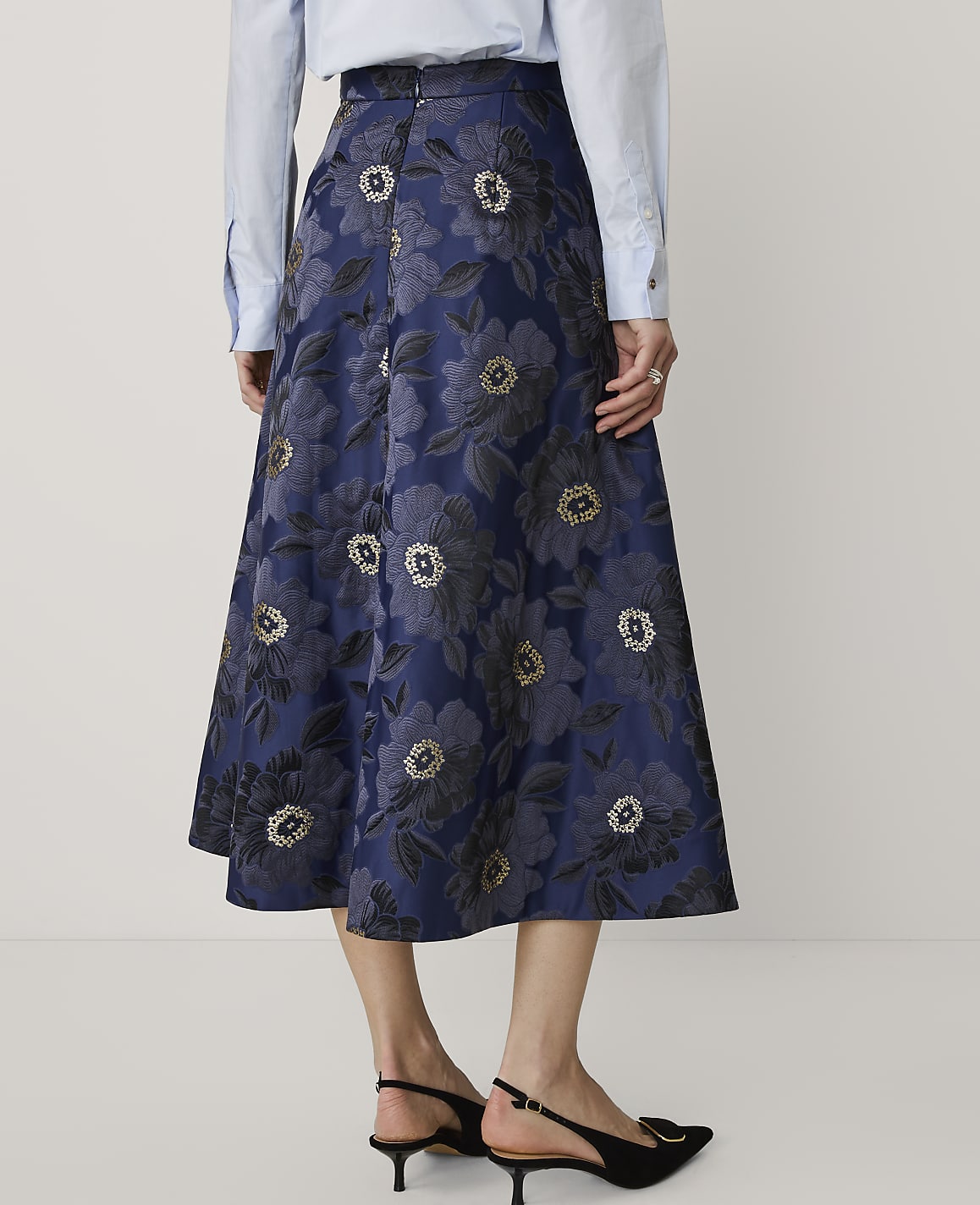A person wears a light blue shirt and a navy blue floral jacquard skirt with black heels against a plain white background.