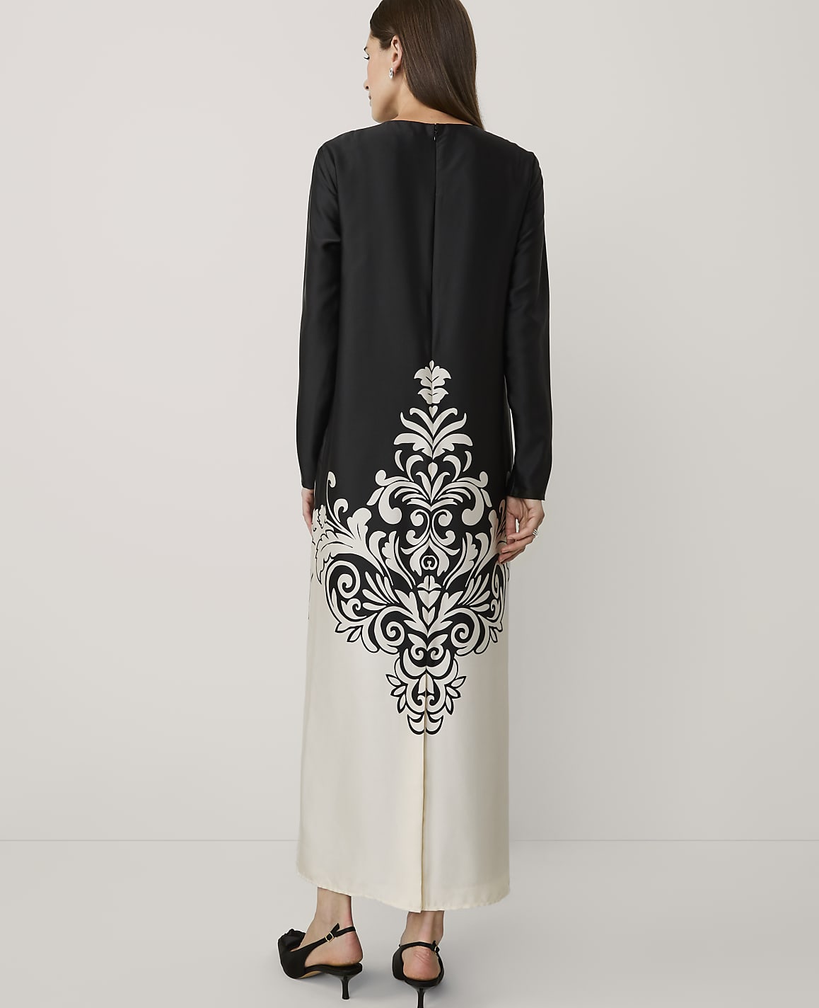 A woman stands with her back to the camera wearing a long black and white gown with a large ornate pattern, set against a plain white background.