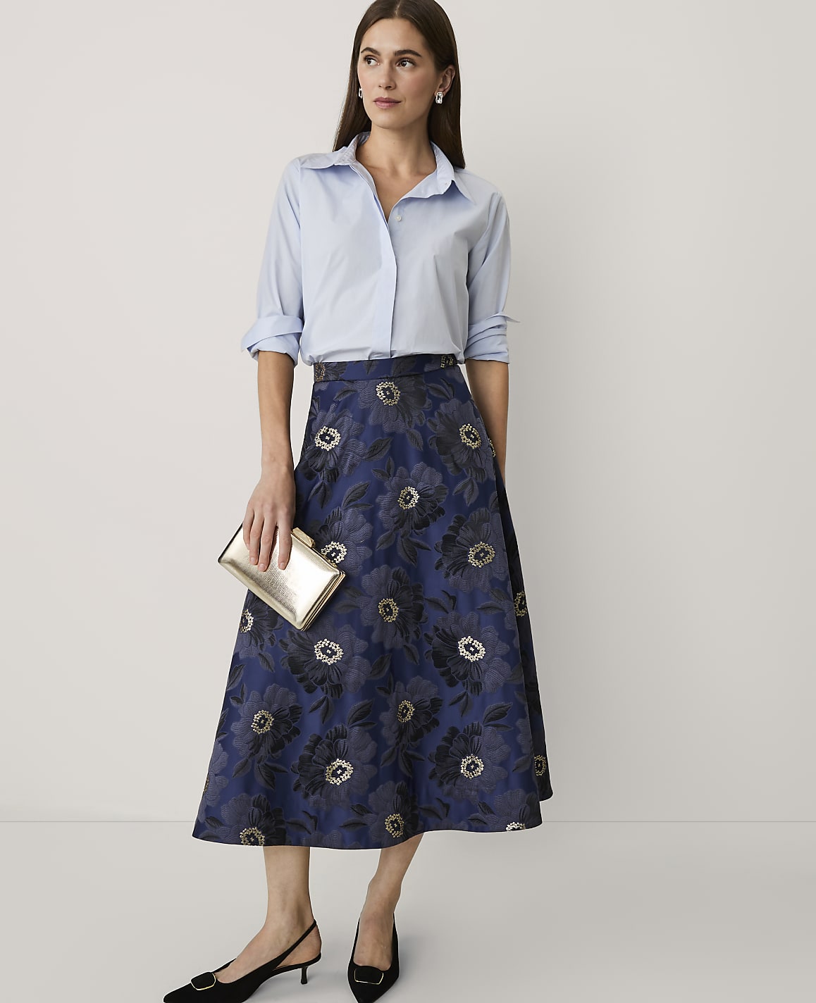 A woman wearing a light blue button-down shirt and a navy floral midi skirt stands against a plain white background.