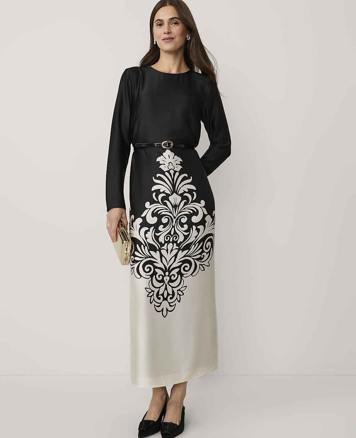 A woman wearing a black and white patterned dress and black heels stands against a plain white background.