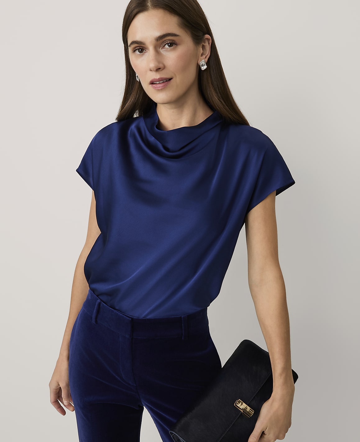 A woman wearing a navy blue satin top and velvet trousers holds a black clutch against a plain light grey background.