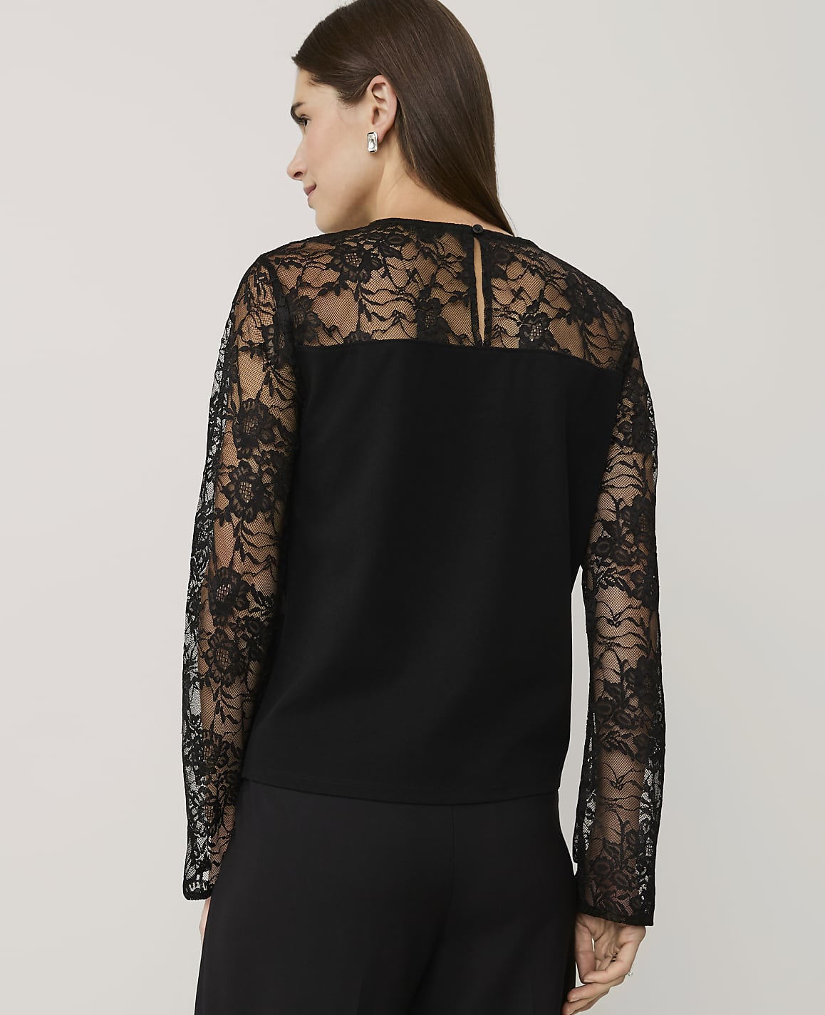 A woman wearing a black lace-detailed top and trousers stands against a plain, light-colored background.