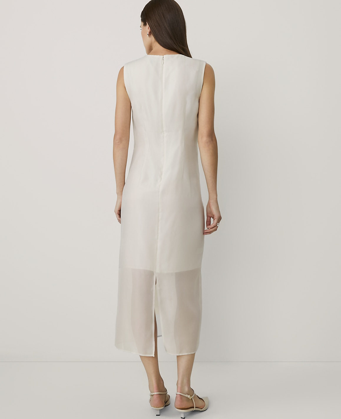 A woman stands with her back to the camera wearing a sleeveless white midi dress against a plain white background.