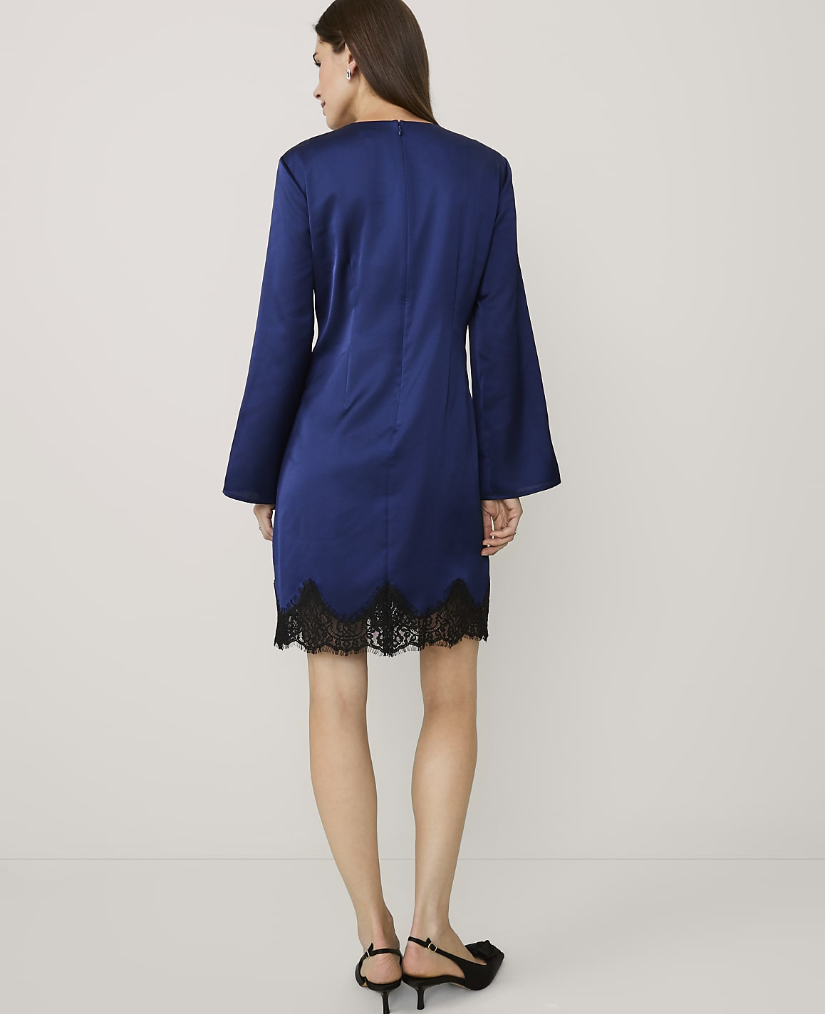 A woman wearing a royal blue dress with black lace trim and black heels stands against a plain white background.