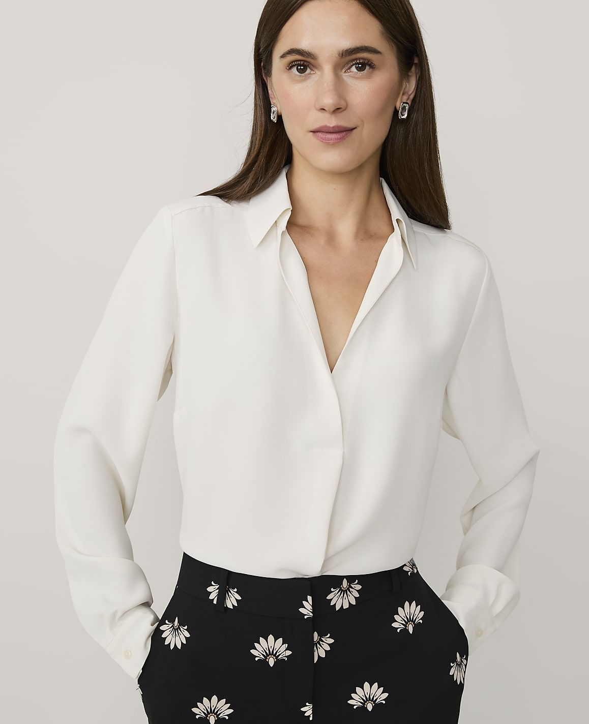 A woman wearing a white long-sleeved blouse and black floral trousers stands against a plain light gray background.