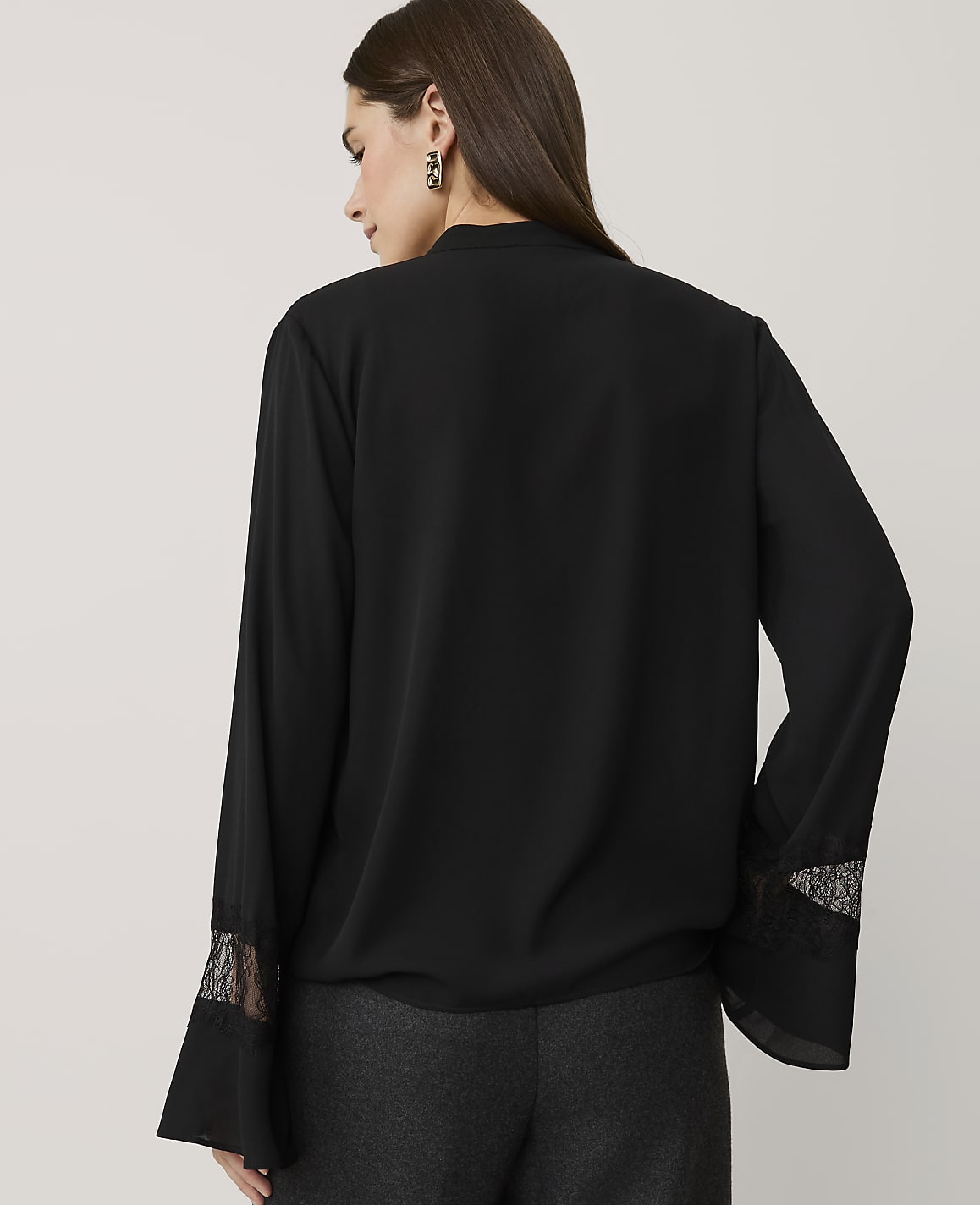 A woman wearing a black long-sleeved blouse with lace cuffs and dark grey trousers stands against a plain, light-colored background.