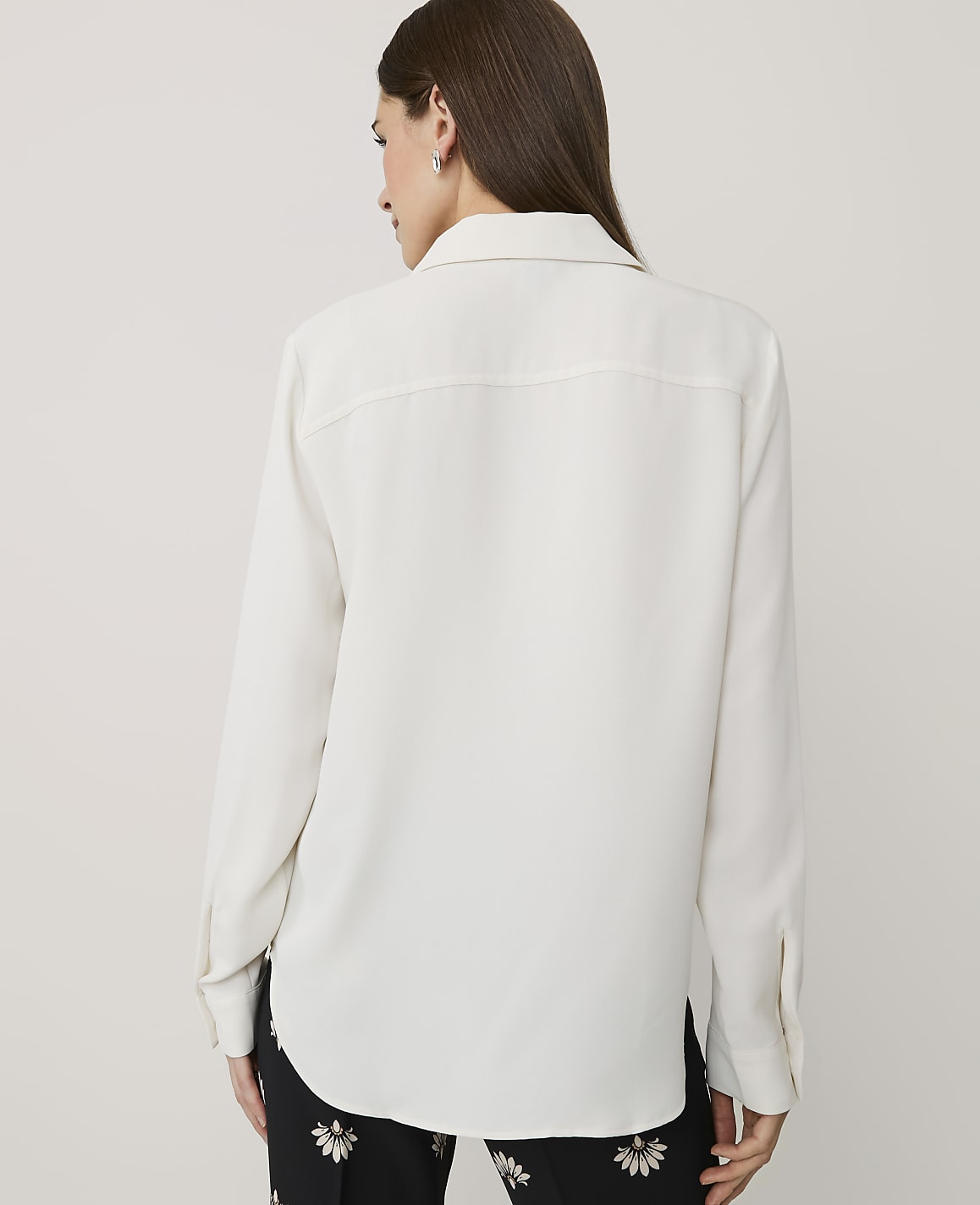 A woman wearing a white long-sleeved shirt and floral black pants stands against a plain light gray background.