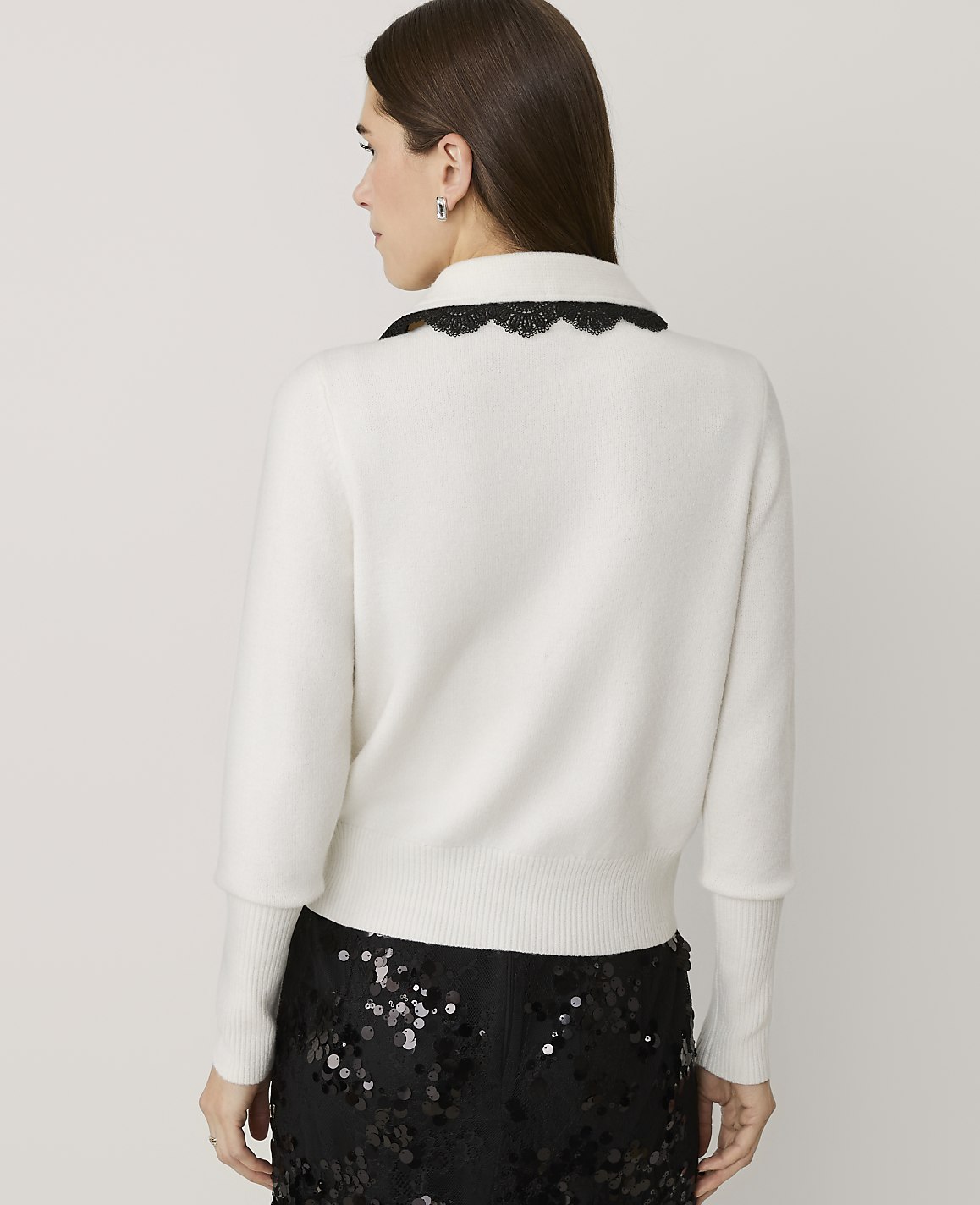 A woman wearing a white sweater with a black lace collar and sequined black pants stands against a plain light gray background.