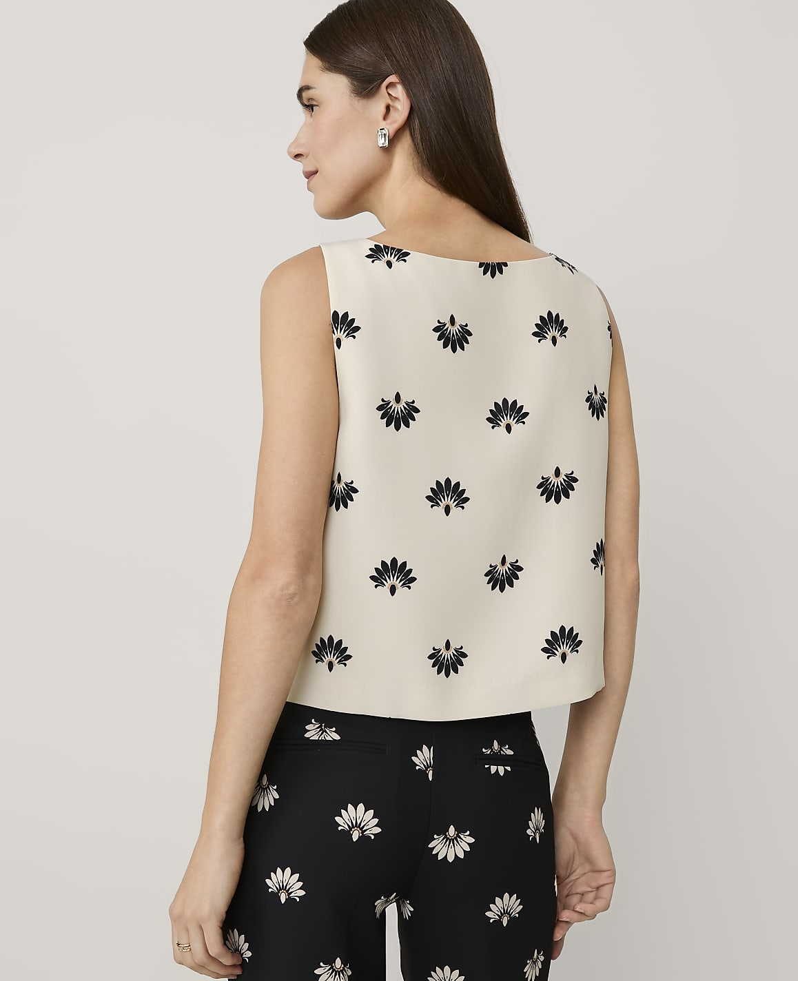A woman stands with her back to the camera wearing a cream floral print top and black floral print pants against a plain light gray background.