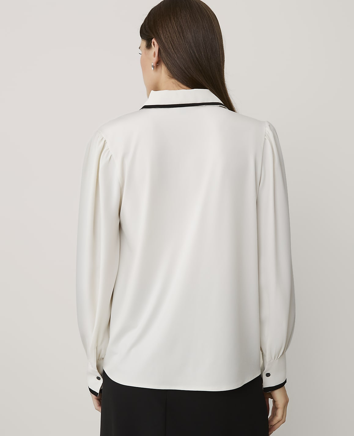 A woman wearing a white long-sleeved blouse and black trousers stands with her back to the camera against a plain light grey background.