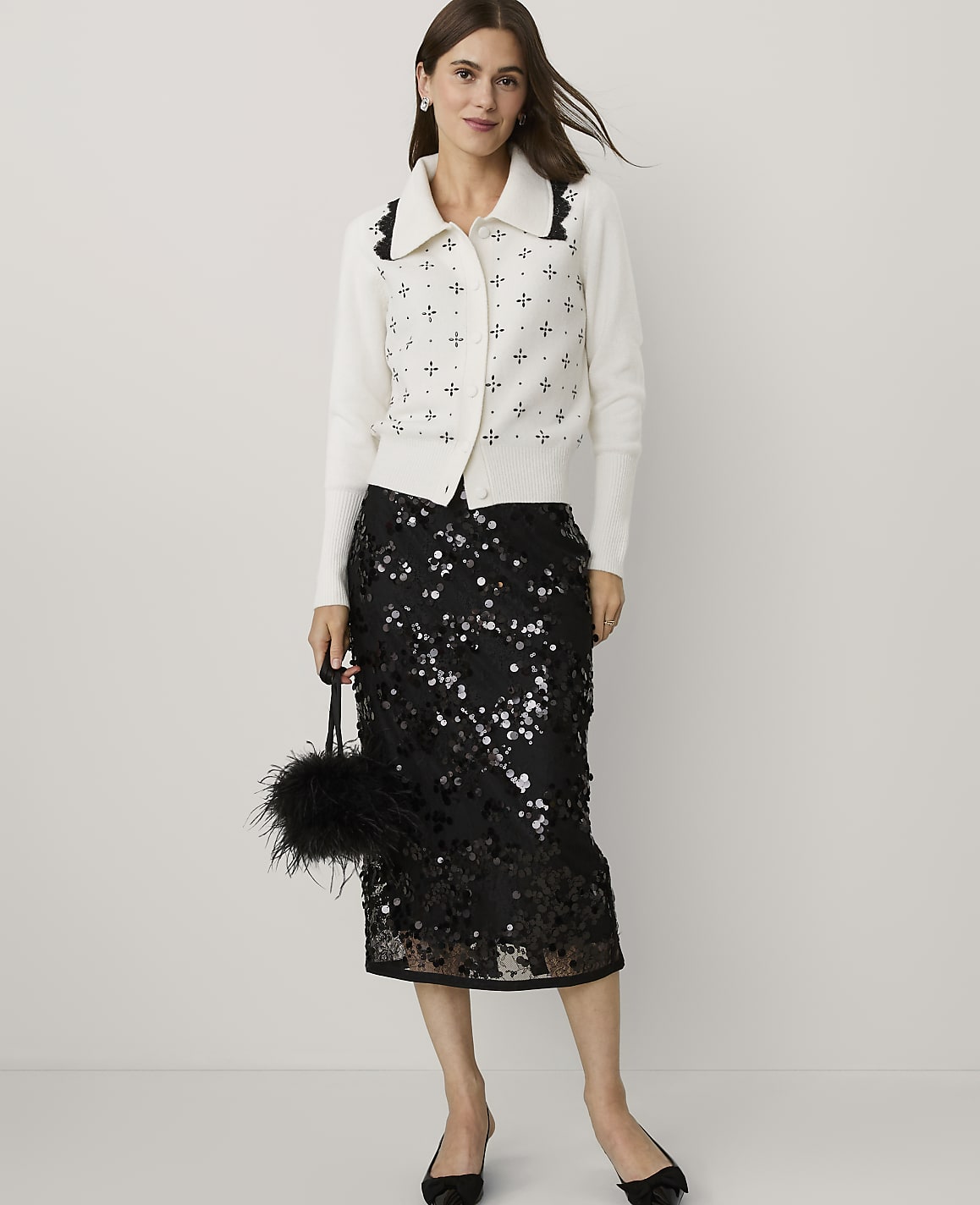 A woman wearing a white patterned cardigan and a black sequined skirt stands against a plain light gray background.