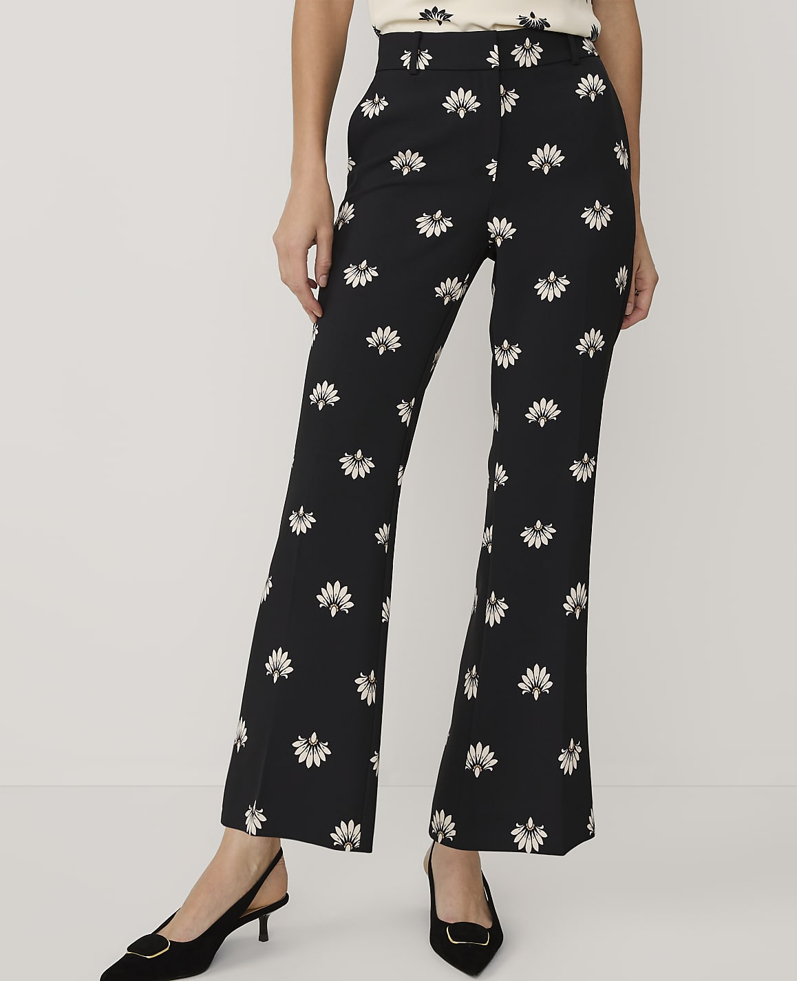 A person wears black floral-print trousers and black pointed-toe heels against a plain white background.