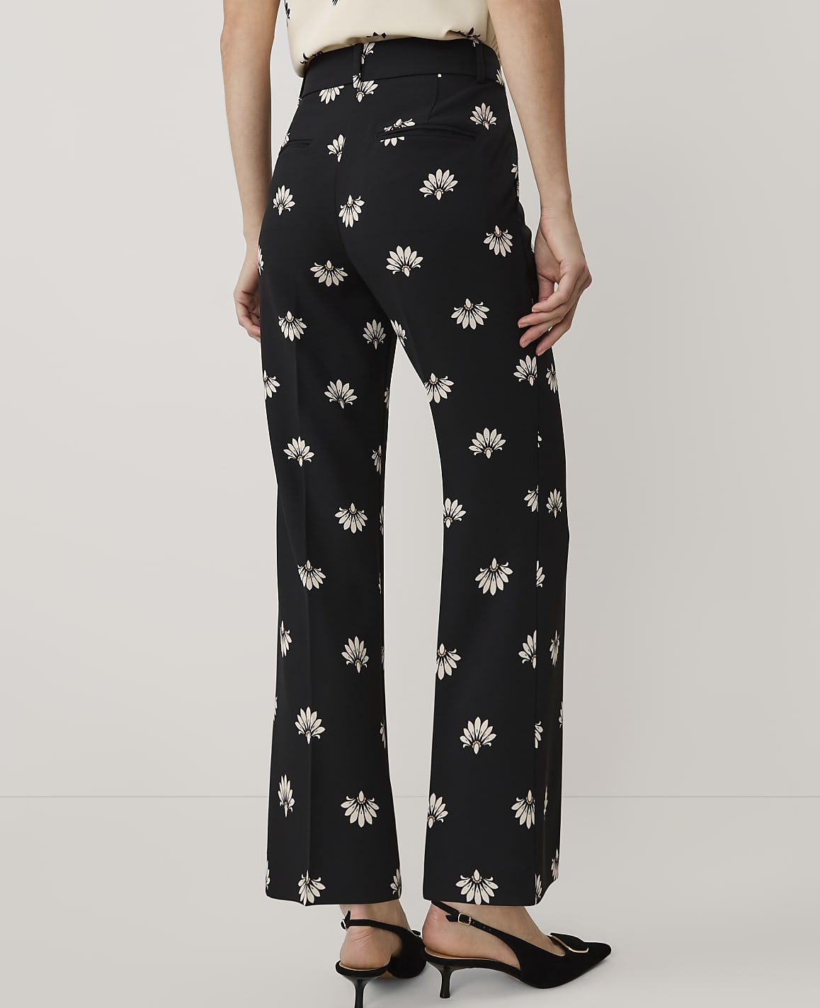 A person stands facing away from the camera wearing black floral trousers and black heels against a plain light grey background.