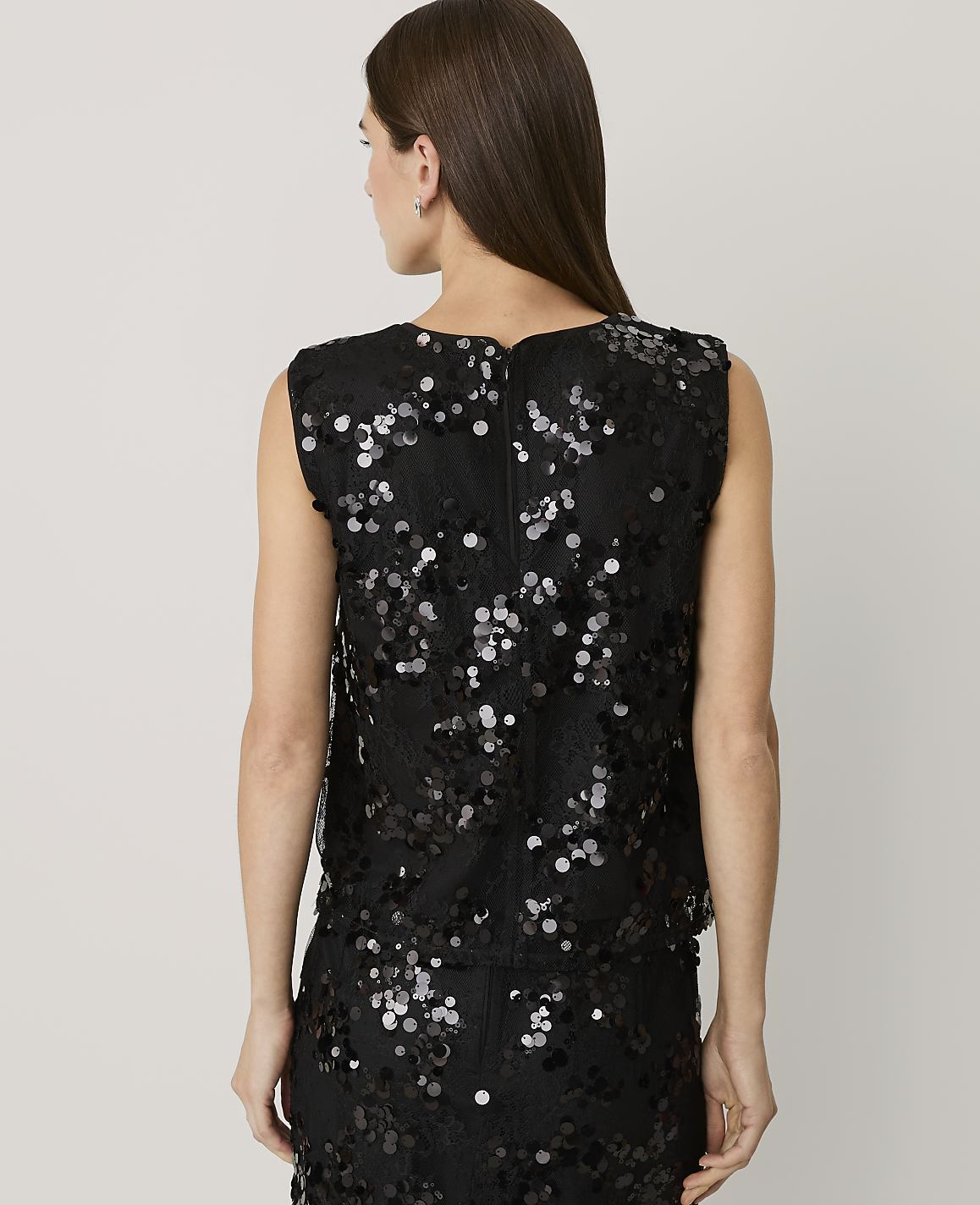 A woman with long brown hair stands with her back to the camera wearing a black sequined top and skirt against a plain light gray background.