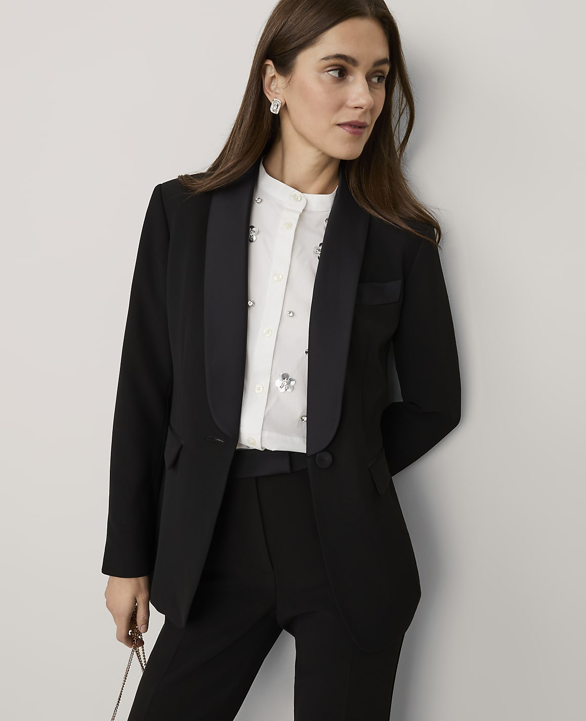 A woman wearing a black blazer, white embellished shirt, and black trousers stands against a plain, light-colored wall.