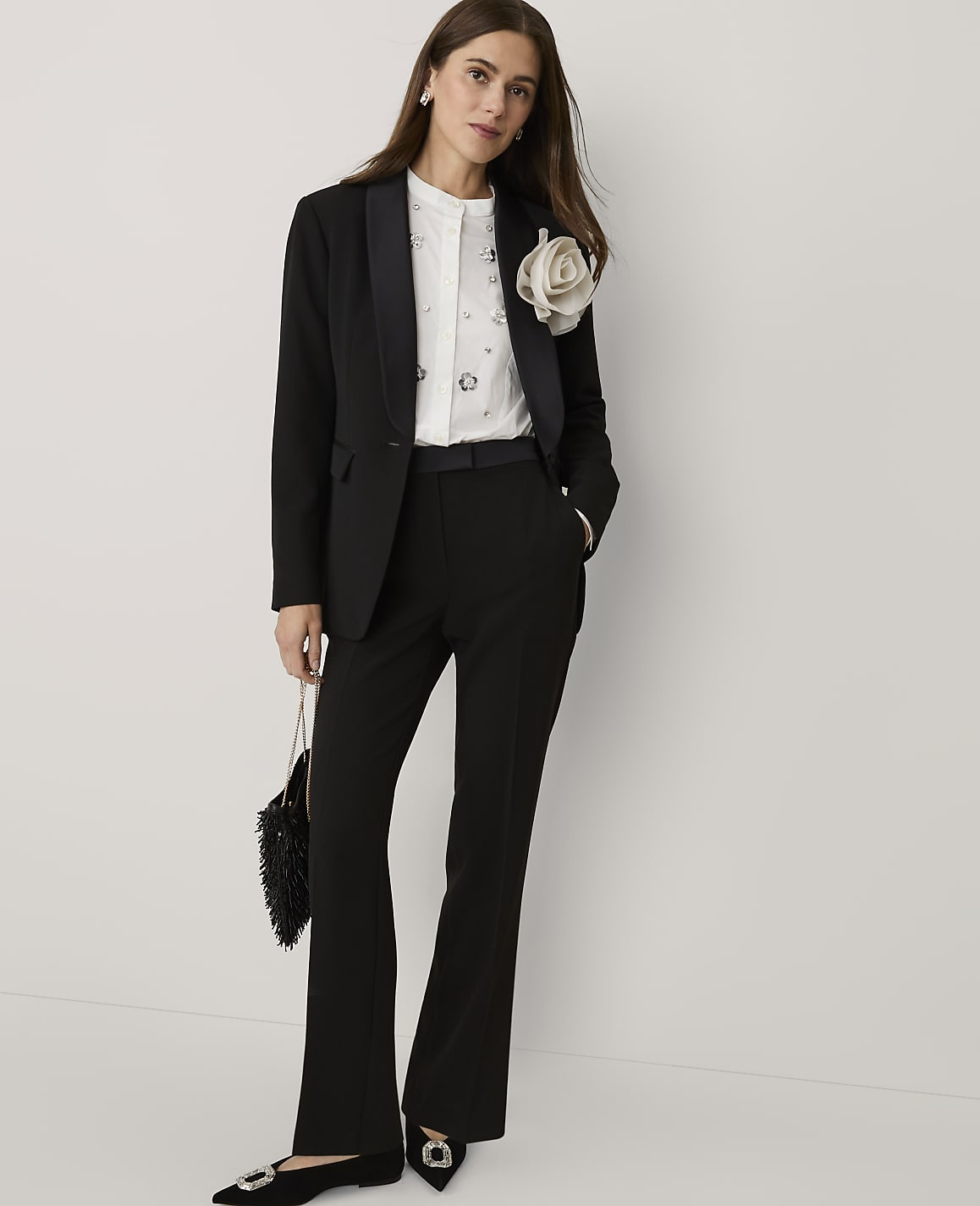 A woman wearing a black suit with a white embellished shirt and a large floral brooch stands against a plain white background.