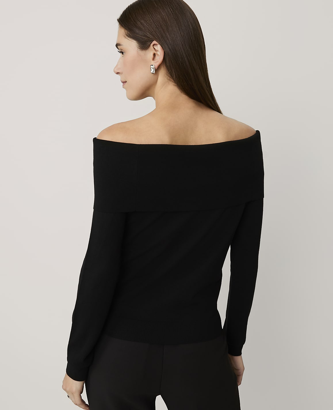 A woman wearing a black off-the-shoulder top and black trousers stands against a plain, light-colored background.