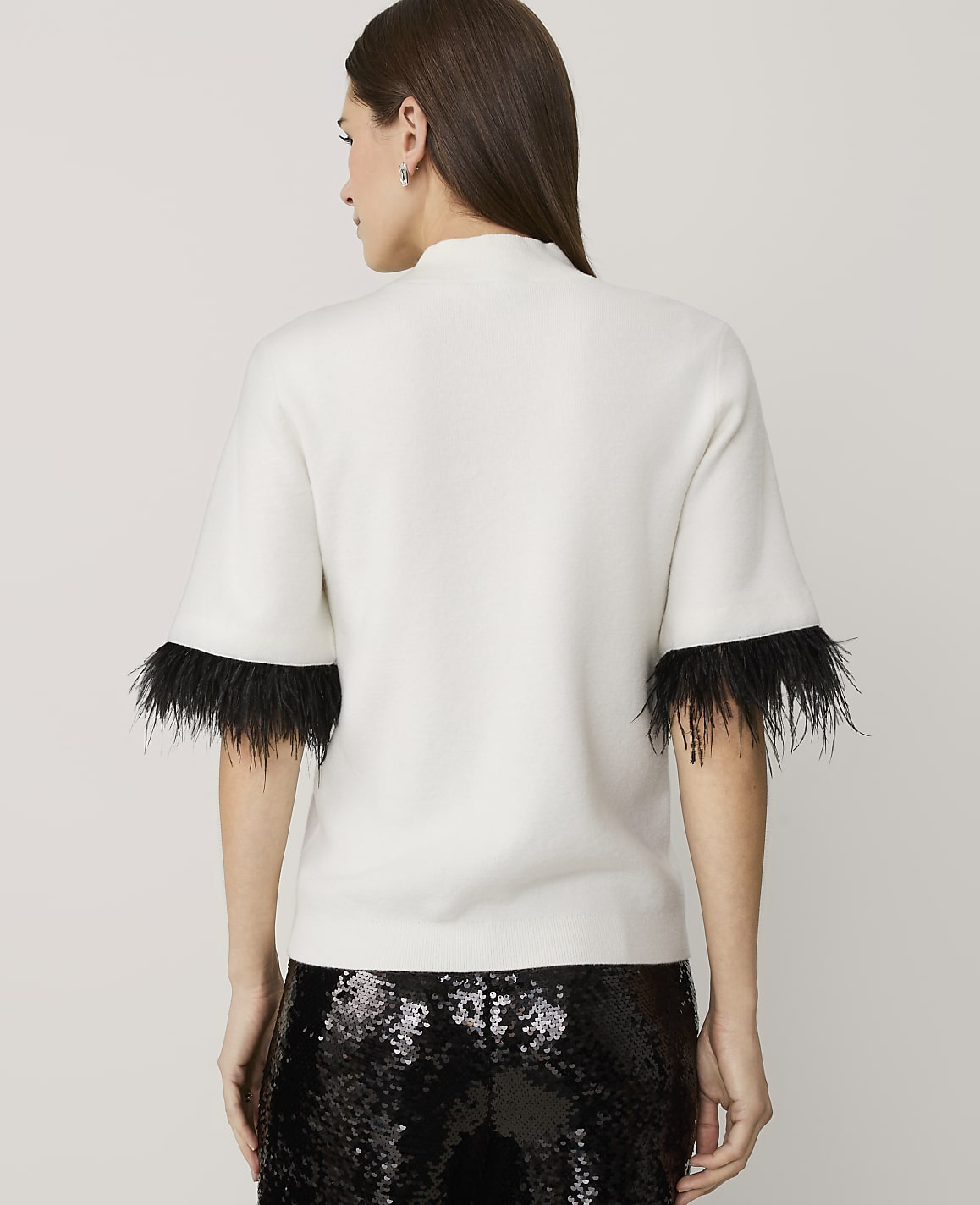 A woman stands with her back to the camera wearing a white short-sleeved top with black feather trim and sequined black pants against a plain light grey background.