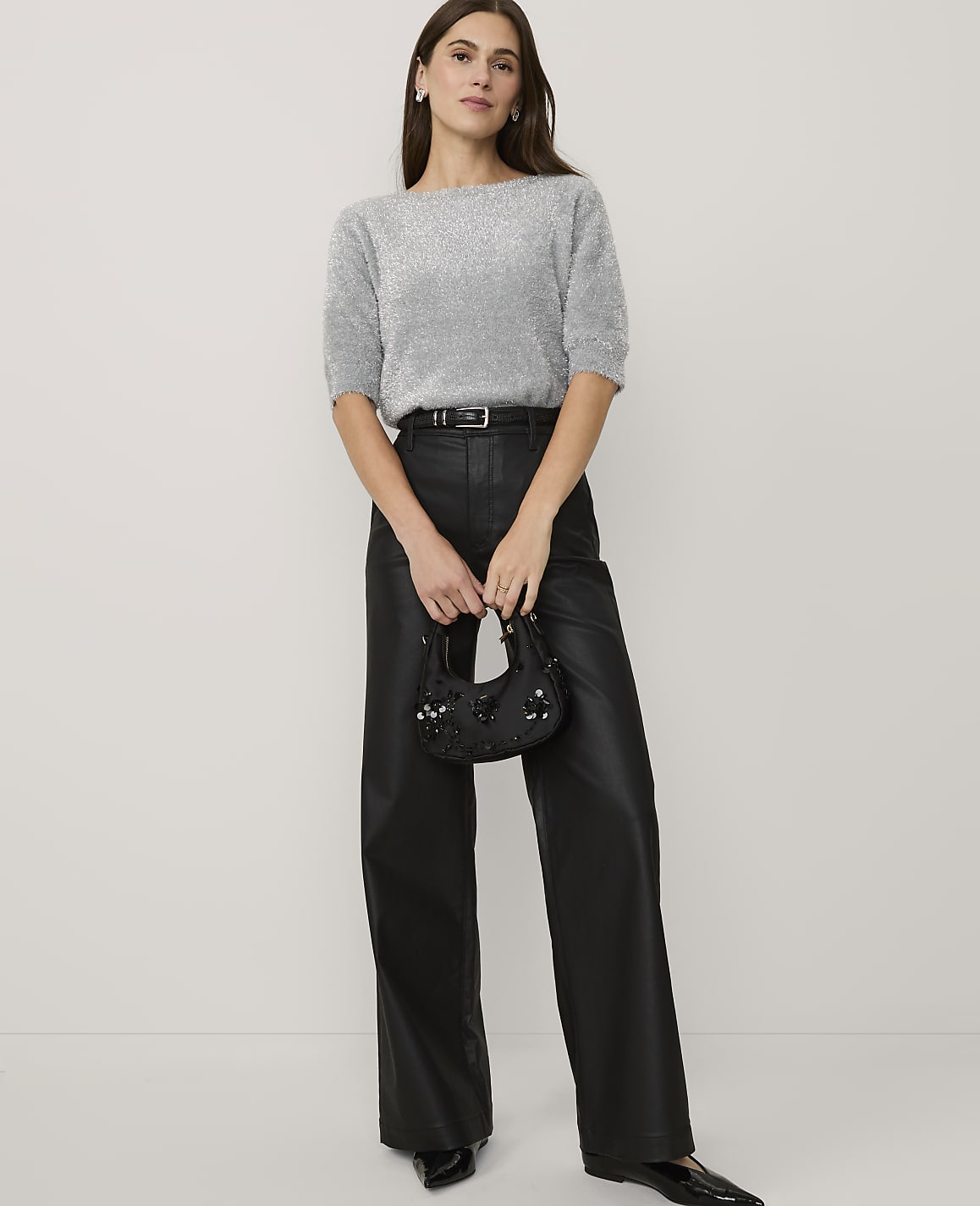 A woman wearing a shimmering silver top and black faux-leather trousers stands against a plain white background.