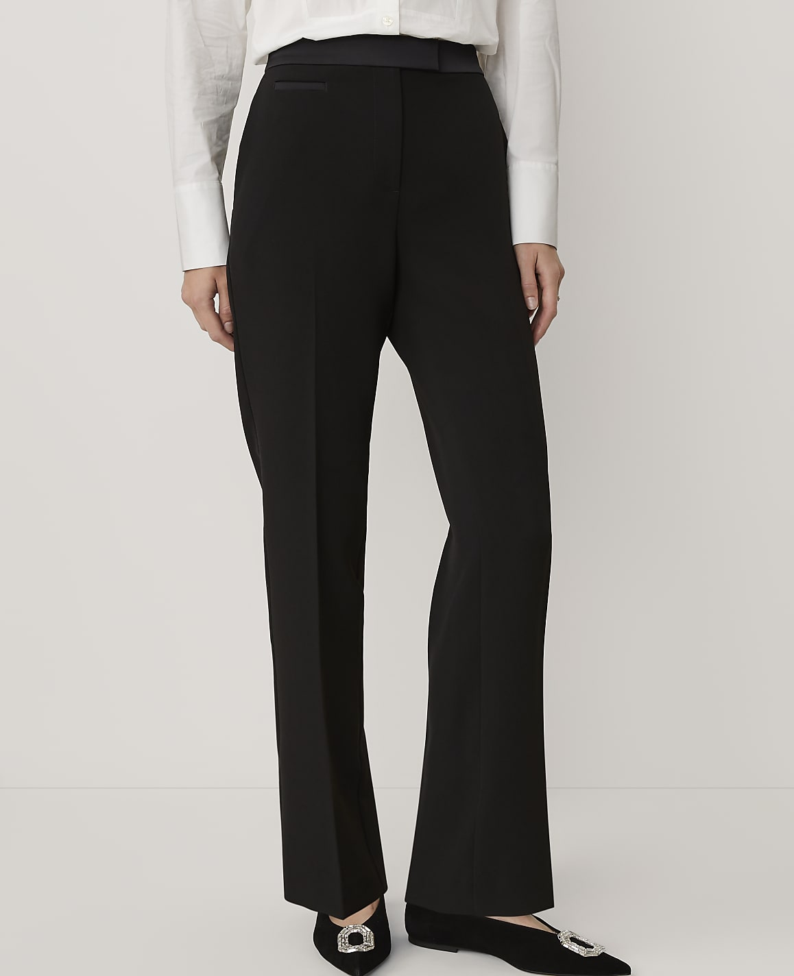 A person wears a white button-down shirt and black trousers with embellished shoes against a plain white background.
