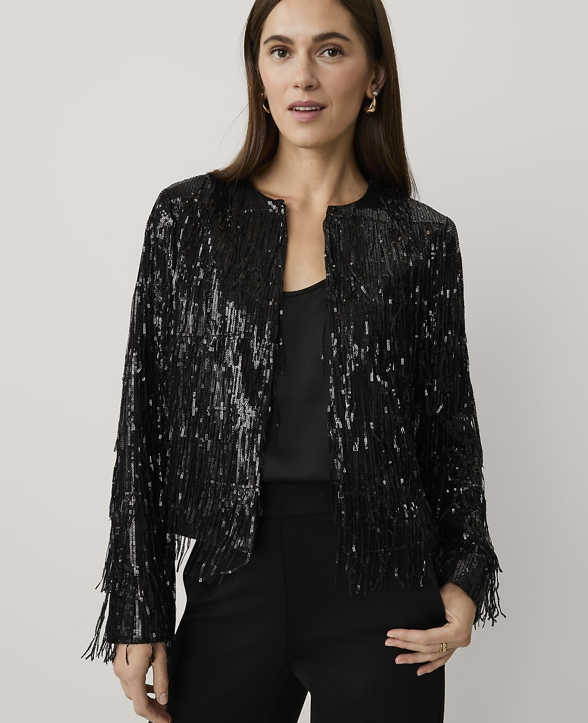 A woman wearing a black sequined fringe jacket and black trousers stands against a plain, light-colored background.
