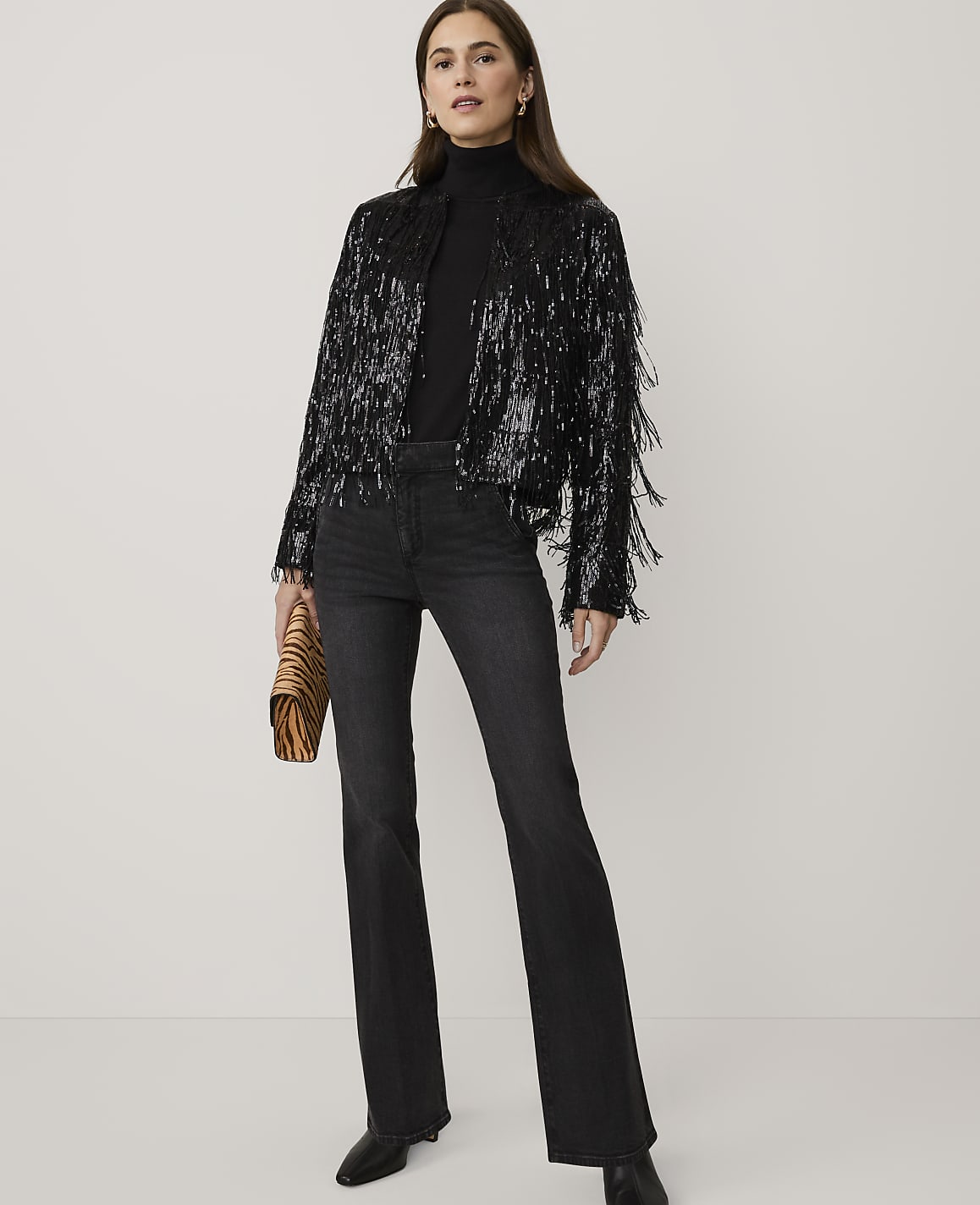 A woman wearing a black sequined fringe jacket and dark flared jeans stands against a plain white background.