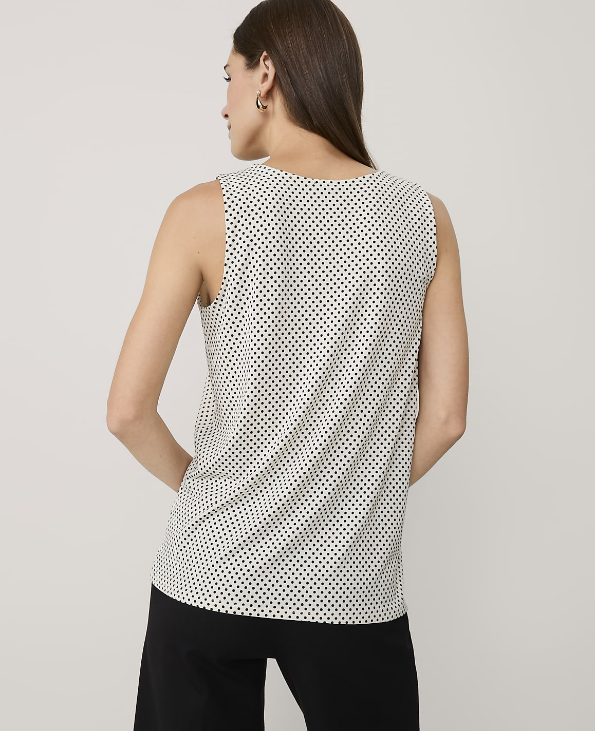 A woman wearing a polka-dot sleeveless top and black pants stands with her back to the camera against a plain light gray background.